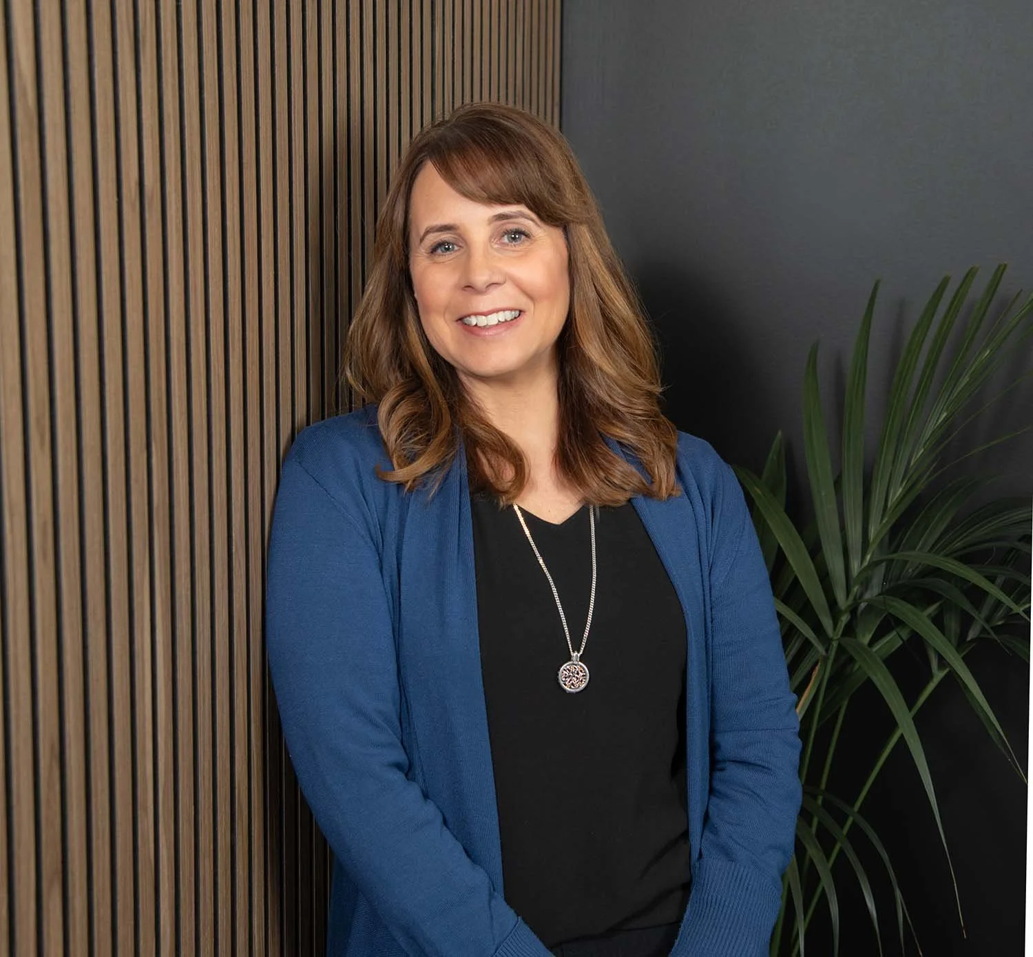 A woman with shoulder-length brown hair, wearing a blue blazer over a black top, standing in front of a wooden wall and next to a green plant, smiling at the camera.