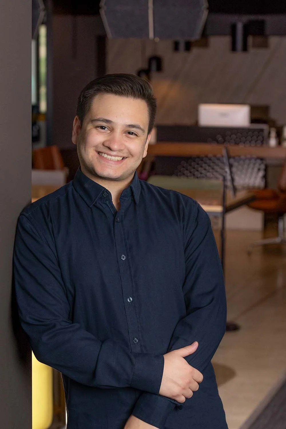 A young man in a dark blue button-up shirt with short dark hair, smiling, standing in a modern indoor setting with wooden elements and seating in the background.