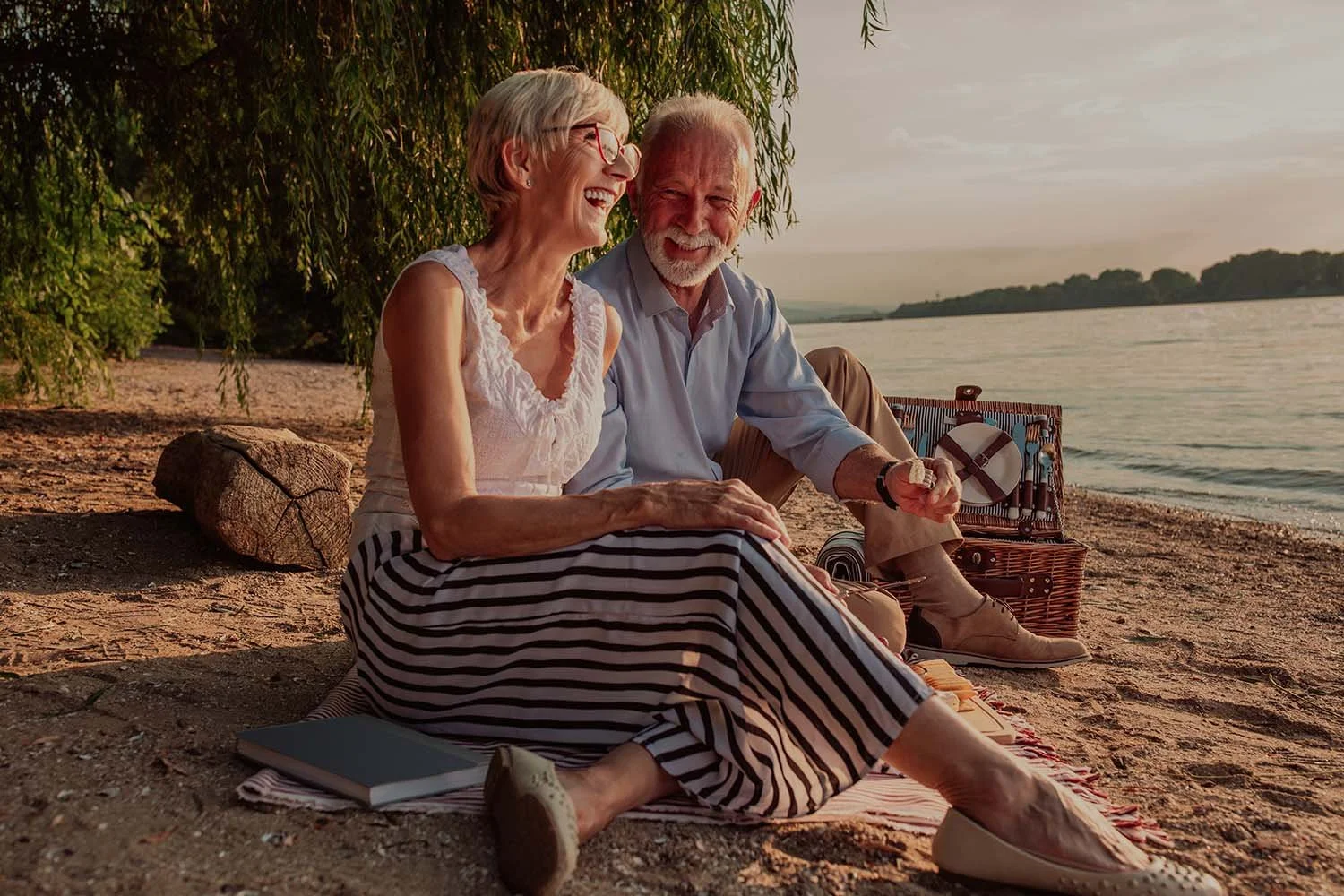 An elderly couple in Adelaide sitting on a beach under a tree at sunset, smiling and enjoying each other's company with a picnic basket nearby.