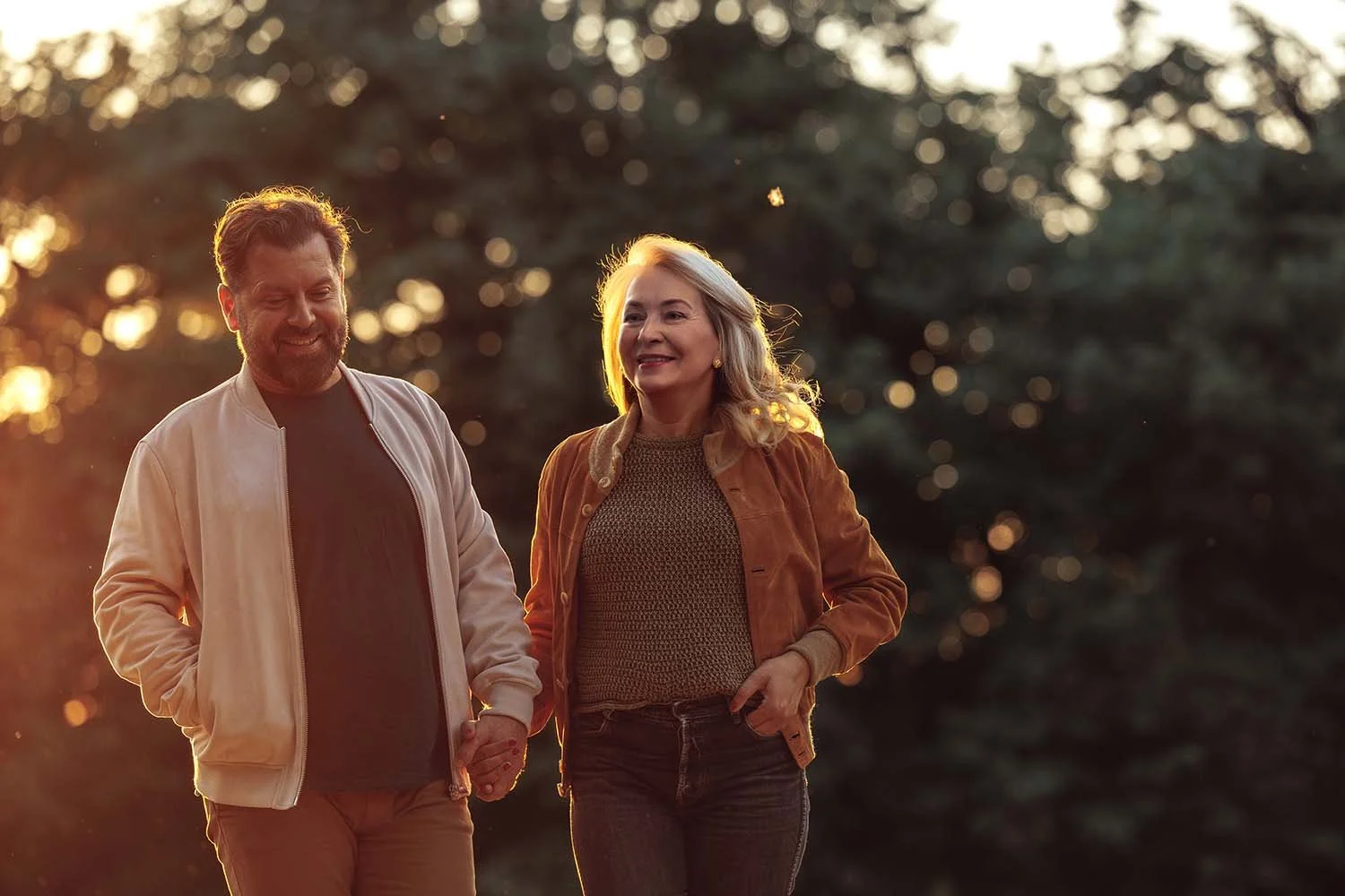 A smiling near retirement couple in Adelaide walking hand in hand outdoors during sunset with trees in the background.