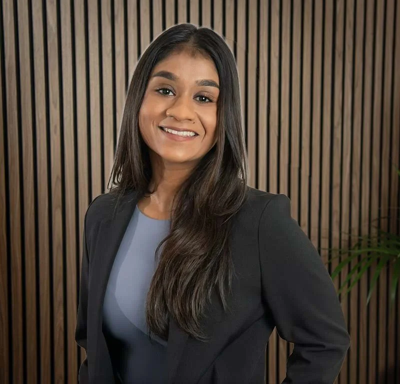 Smiling young woman with long dark hair in a black blazer standing in front of a wooden slat wall.