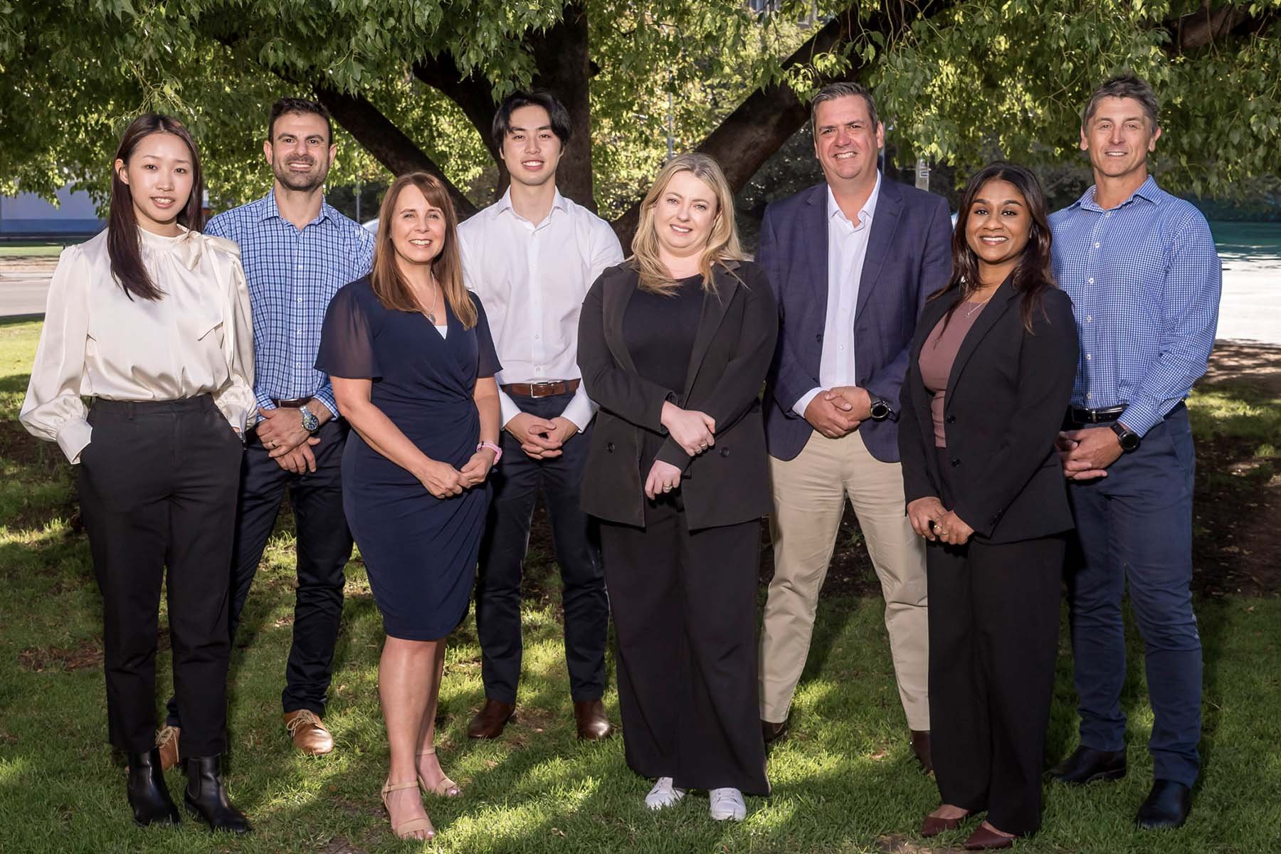Synergy Private Wealth, financial planning for families Adelaide, team photo professionally dressed standing outdoors under a large tree, smiling.