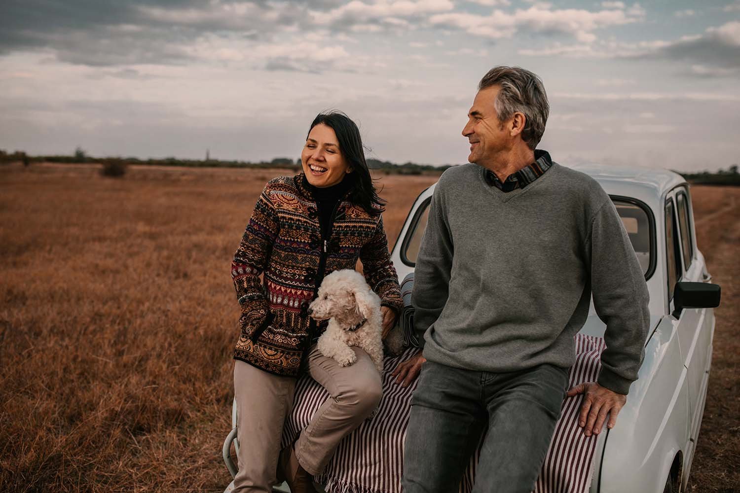 A couple in Adelaide are sitting on the back of a small white vehicle in an open field. The woman is holding a small white dog, and both are smiling at each other. The sky is cloudy and the field has tall, dry grass.