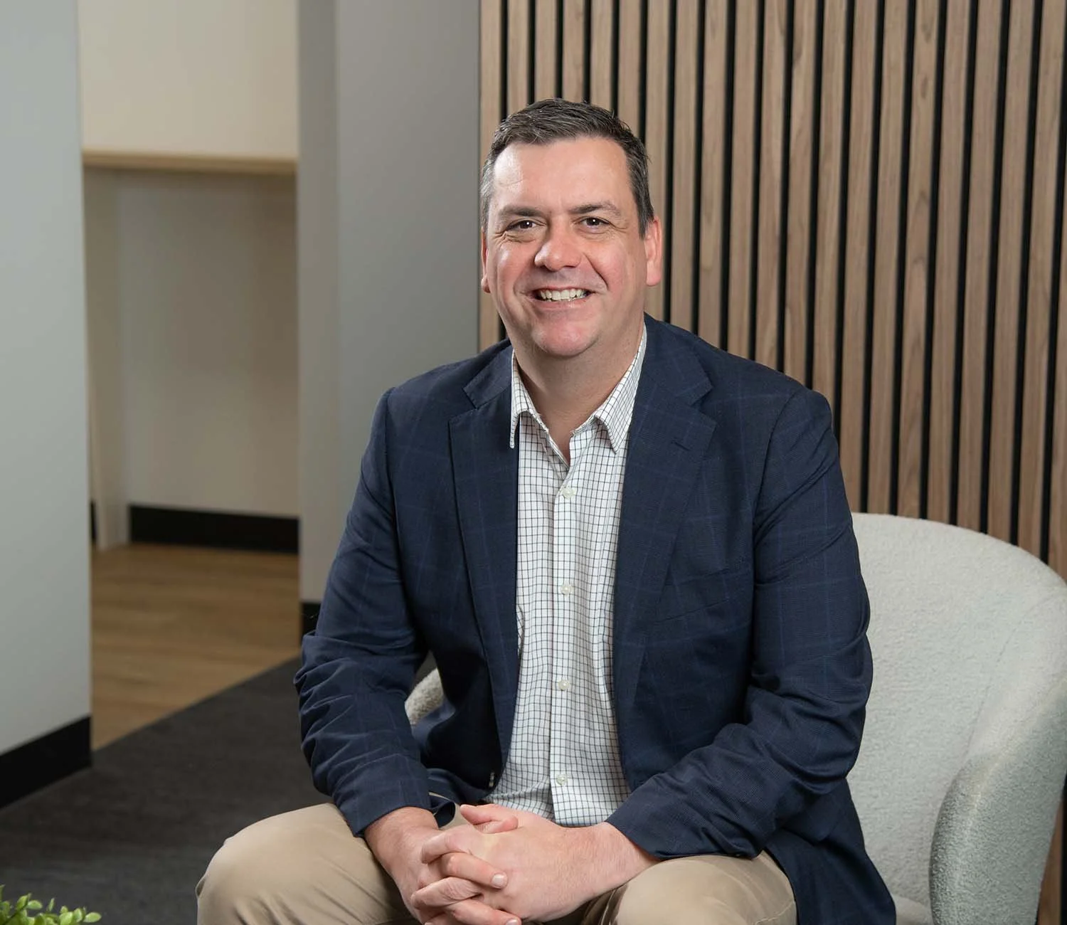 Man sitting on a light-colored chair, wearing a navy blazer and light-colored pants, smiling in a modern office setting with wood panel wall and beige walls.