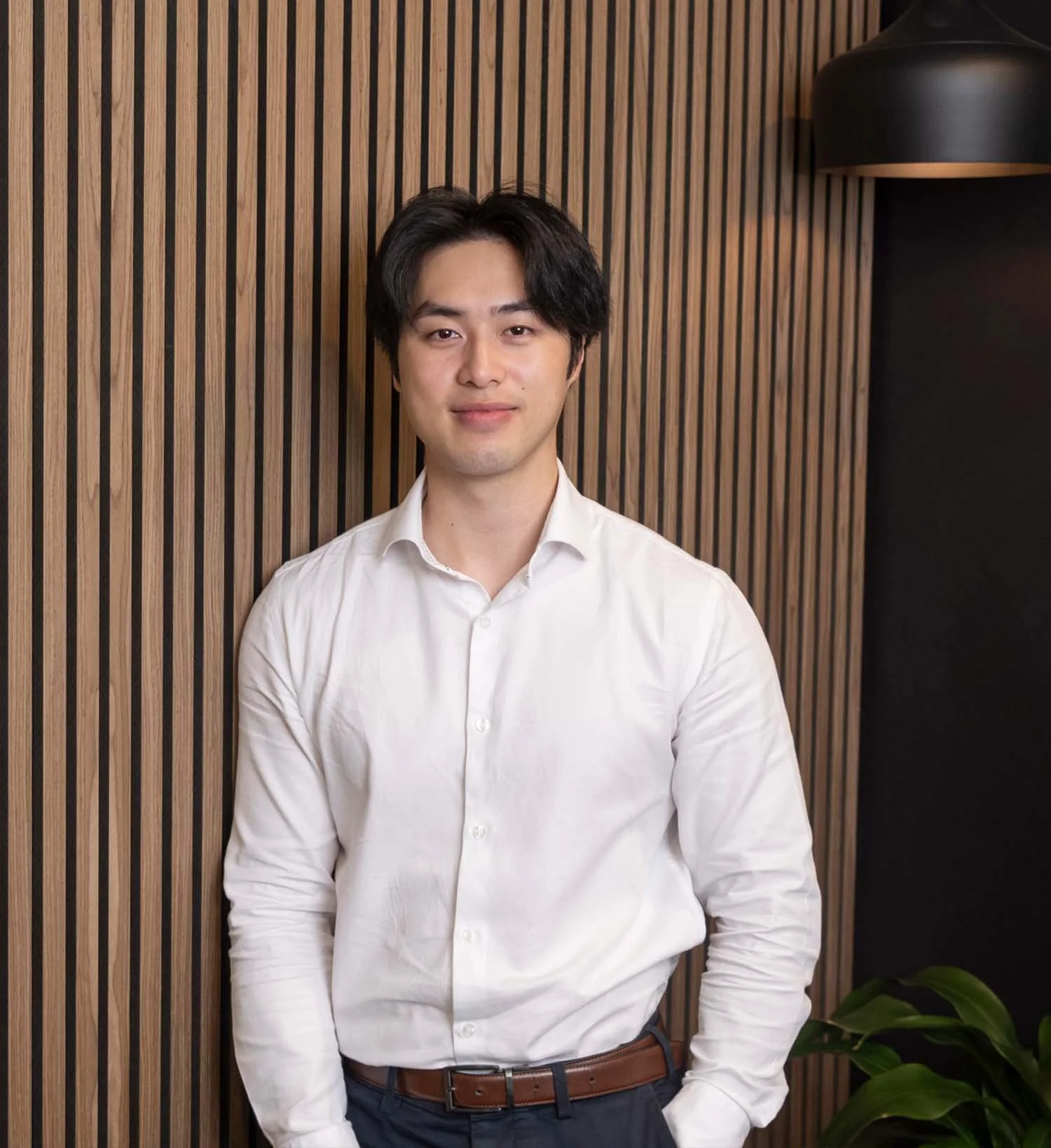 A young man with black hair wearing a white dress shirt and dark pants standing in front of a wooden slat wall with a black lampshade and green plant nearby.