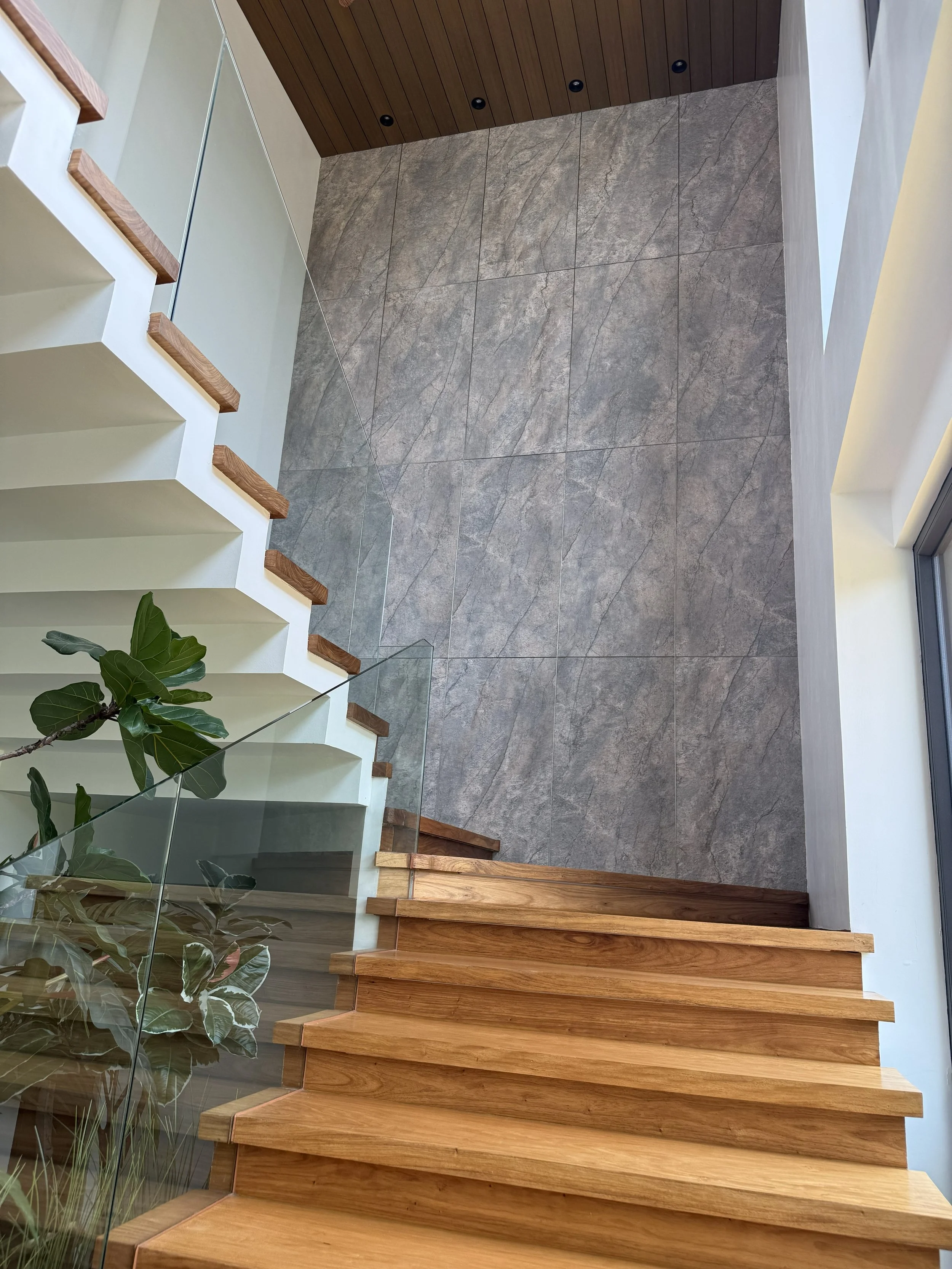 Interior view of a modern staircase with wooden steps, glass railing, large gray tiled floor, a green plant on the lower left, and a wooden ceiling with black recessed lighting.