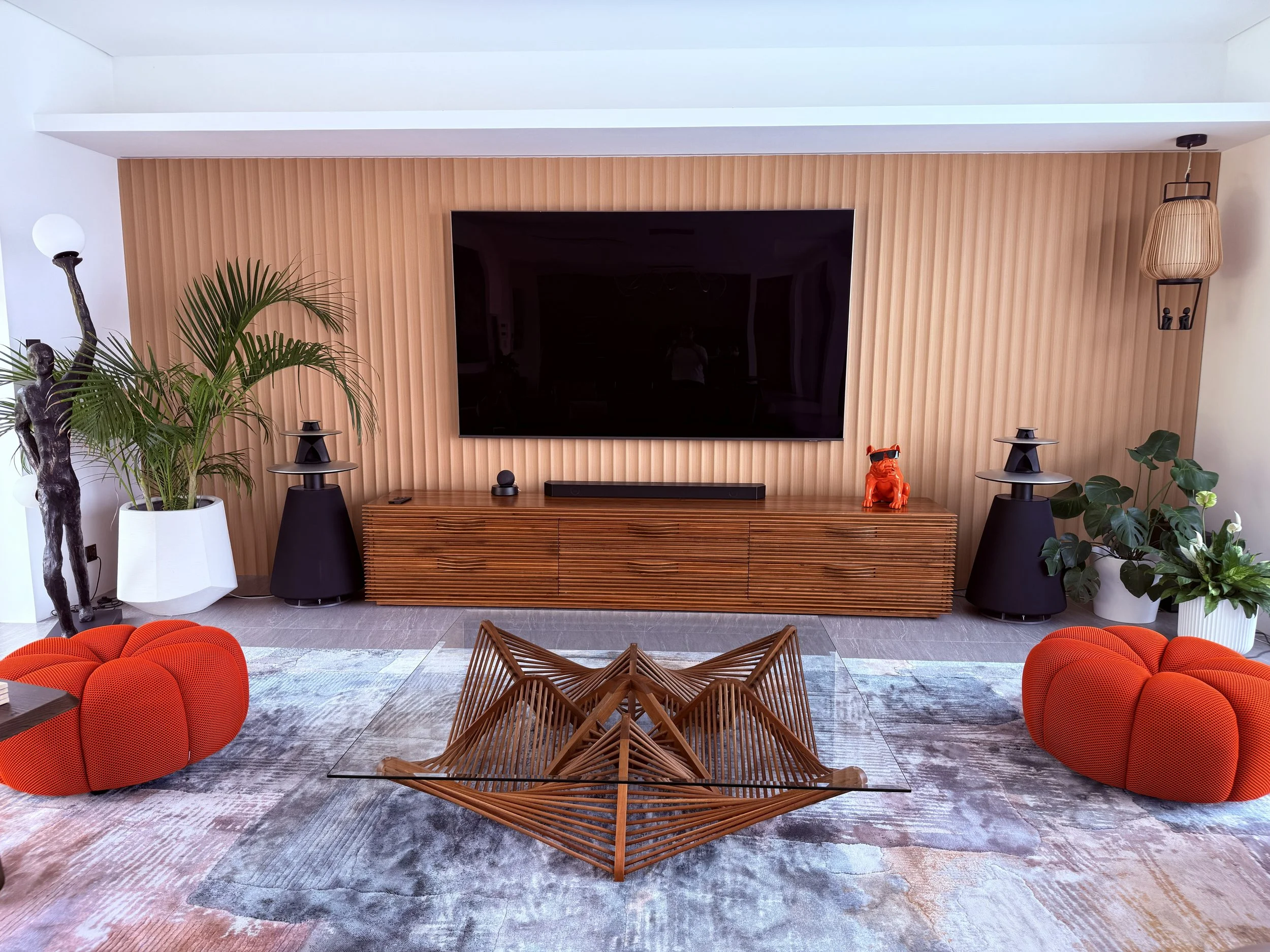 Modern living room with a mounted flat-screen TV on a wooden feature wall, surrounded by plants, unique orange poufs, a glass coffee table with wooden geometric details, a wooden console, and decorative lamps.