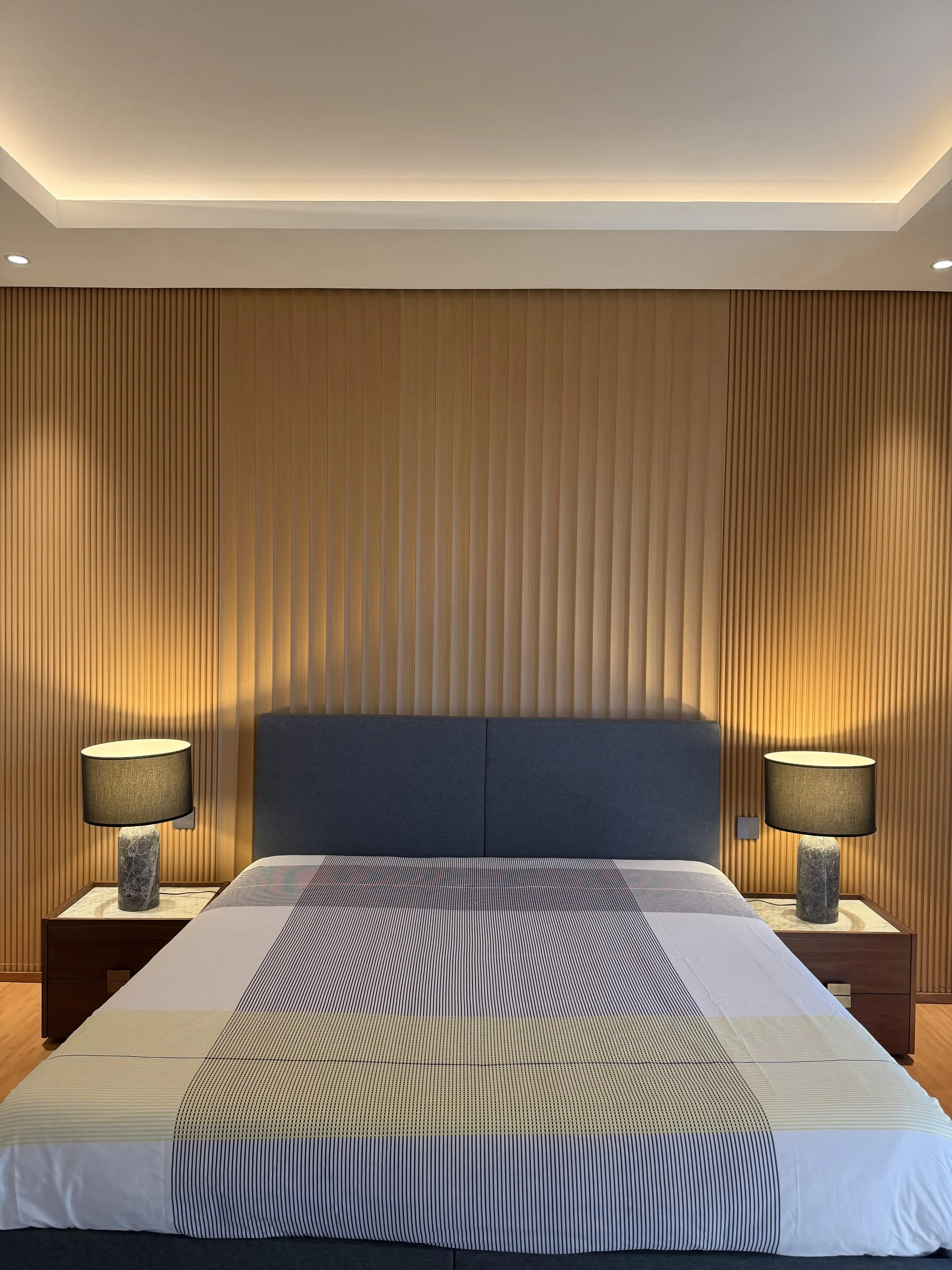 A modern bedroom with a bed and two bedside tables with lamps, wooden wall paneling in the background, and soft ceiling lighting.