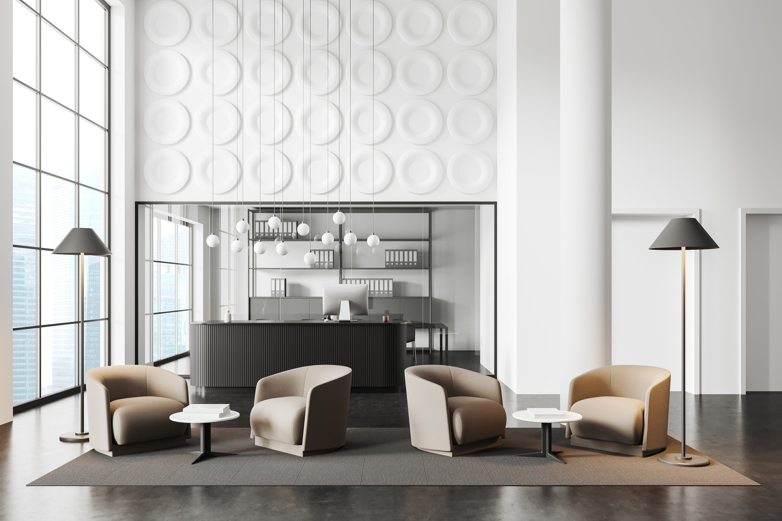 Modern office reception area with beige chairs, small round tables, tall floor lamps, large windows, white walls, and a black reception desk with shelves and binders in the background.