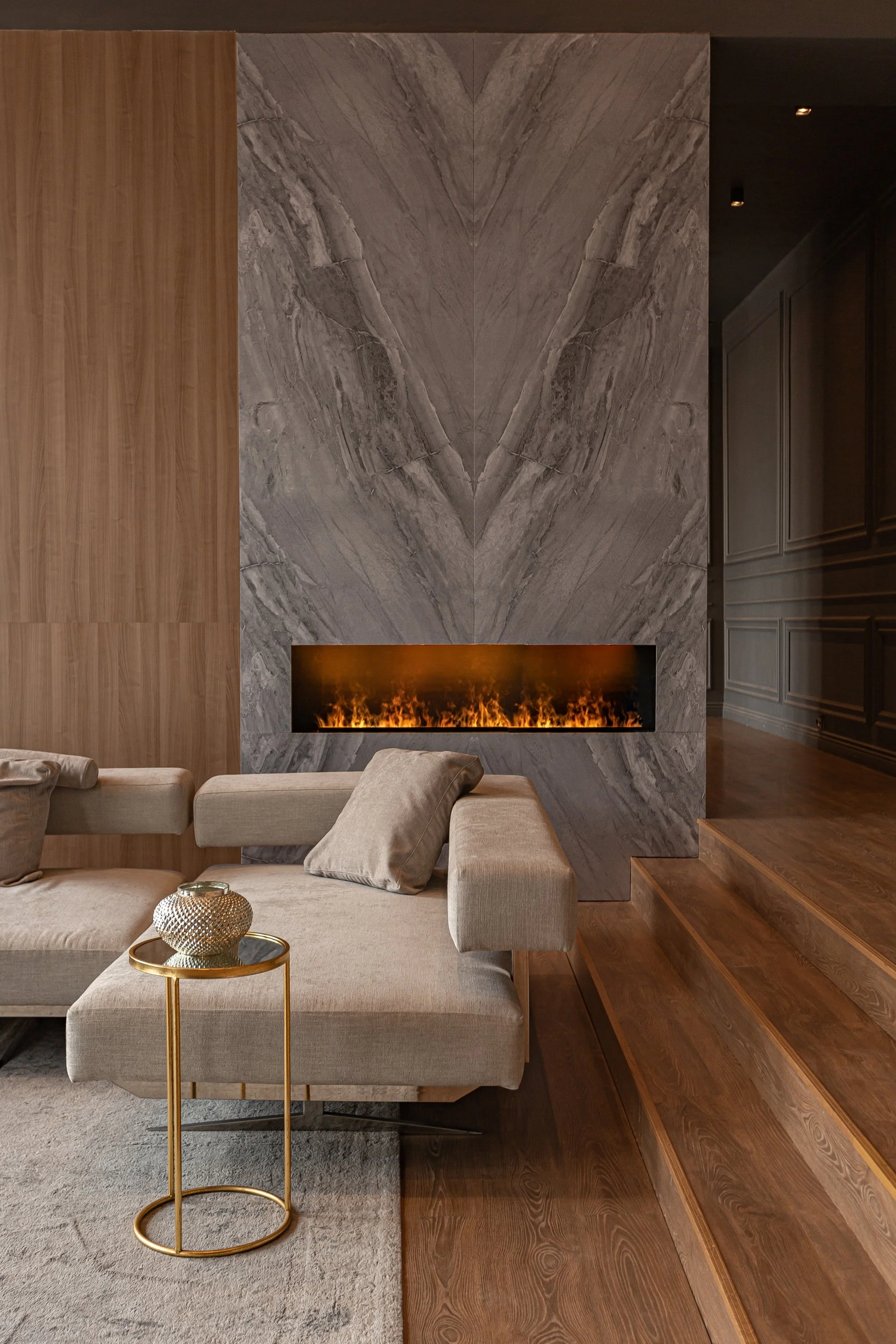 Interior living room with a beige sofa, decorative pillows, a small gold side table with a decorative object, a gray marble fireplace with a glowing fire, wooden floor and steps, and dark wall paneling.
