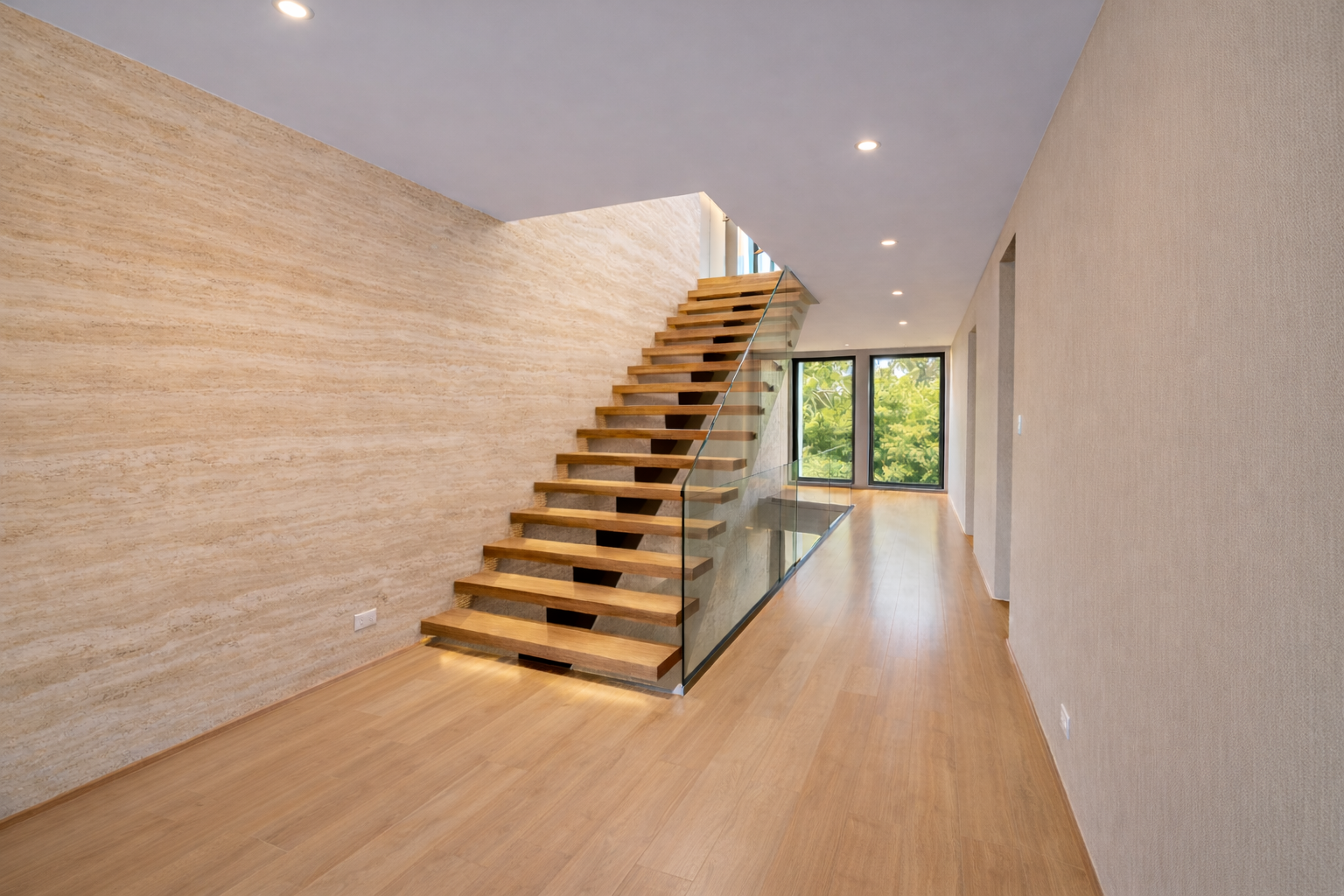 A modern interior with wooden flooring, a floating staircase with wooden steps and glass railing, and large windows showing green trees outside.