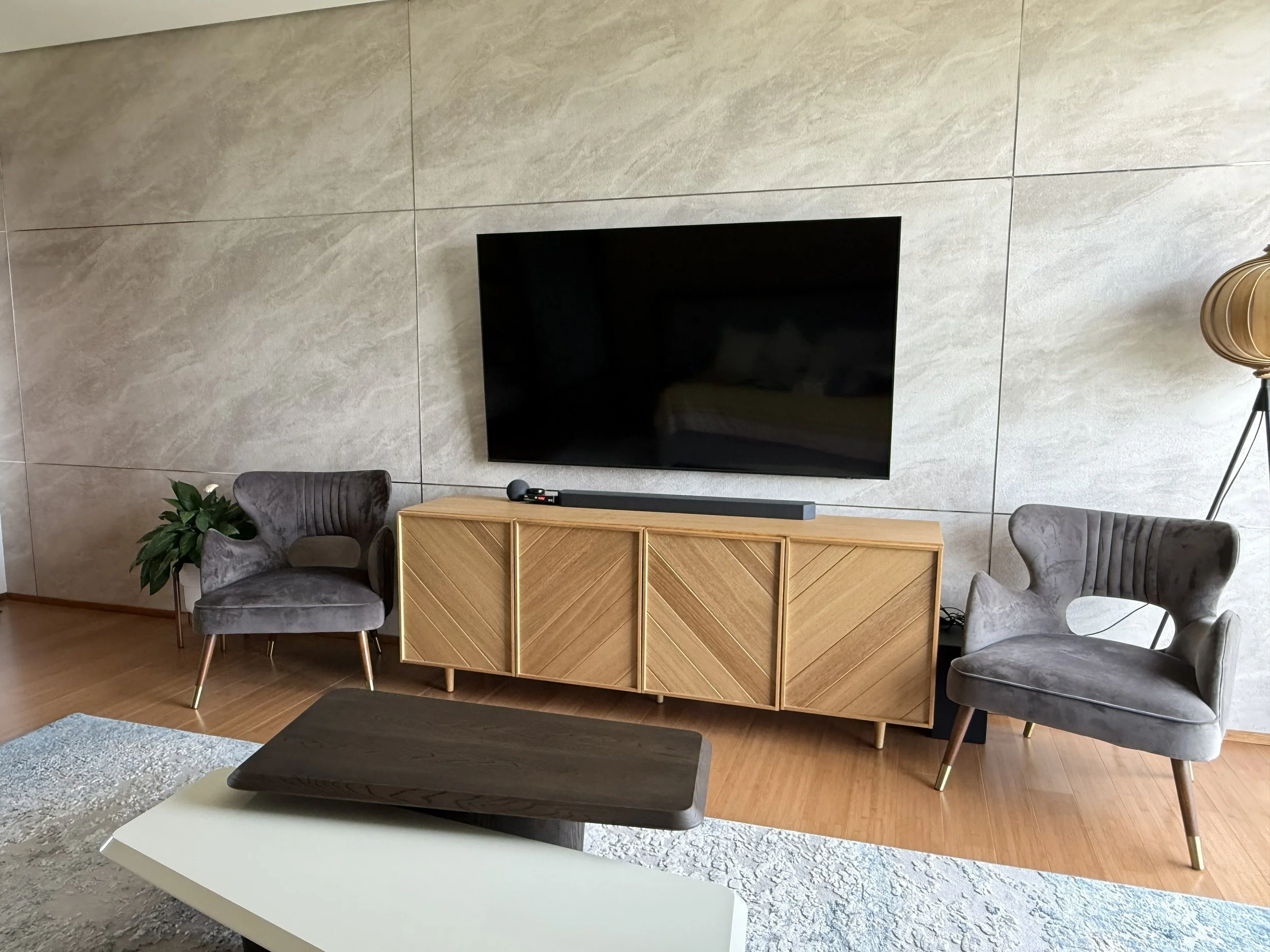 Modern living room with wall-mounted flat-screen TV, wooden media console, two gray velvet armchairs, a potted plant, and a standing lamp, on a light-colored wall and wooden floor.