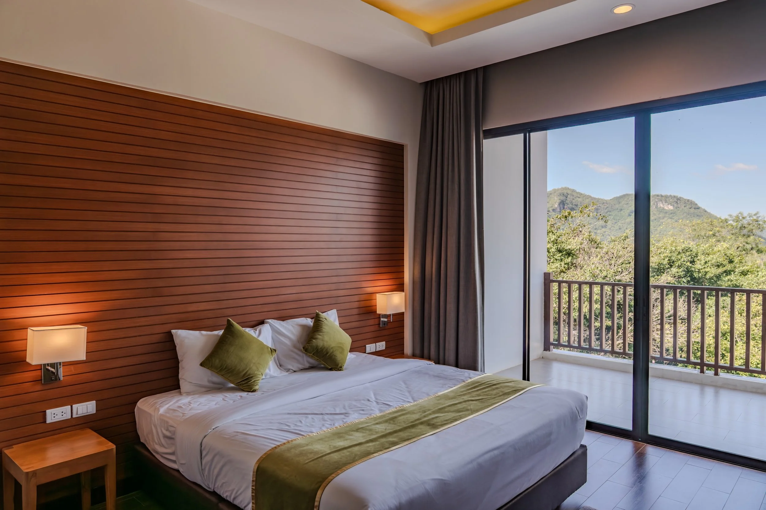 A bedroom with a large bed covered in white linens and green pillows, a wooden headboard with built-in lighting, nightstands on each side with lamps, and a large glass sliding door leading to a balcony overlooking green trees and mountains.