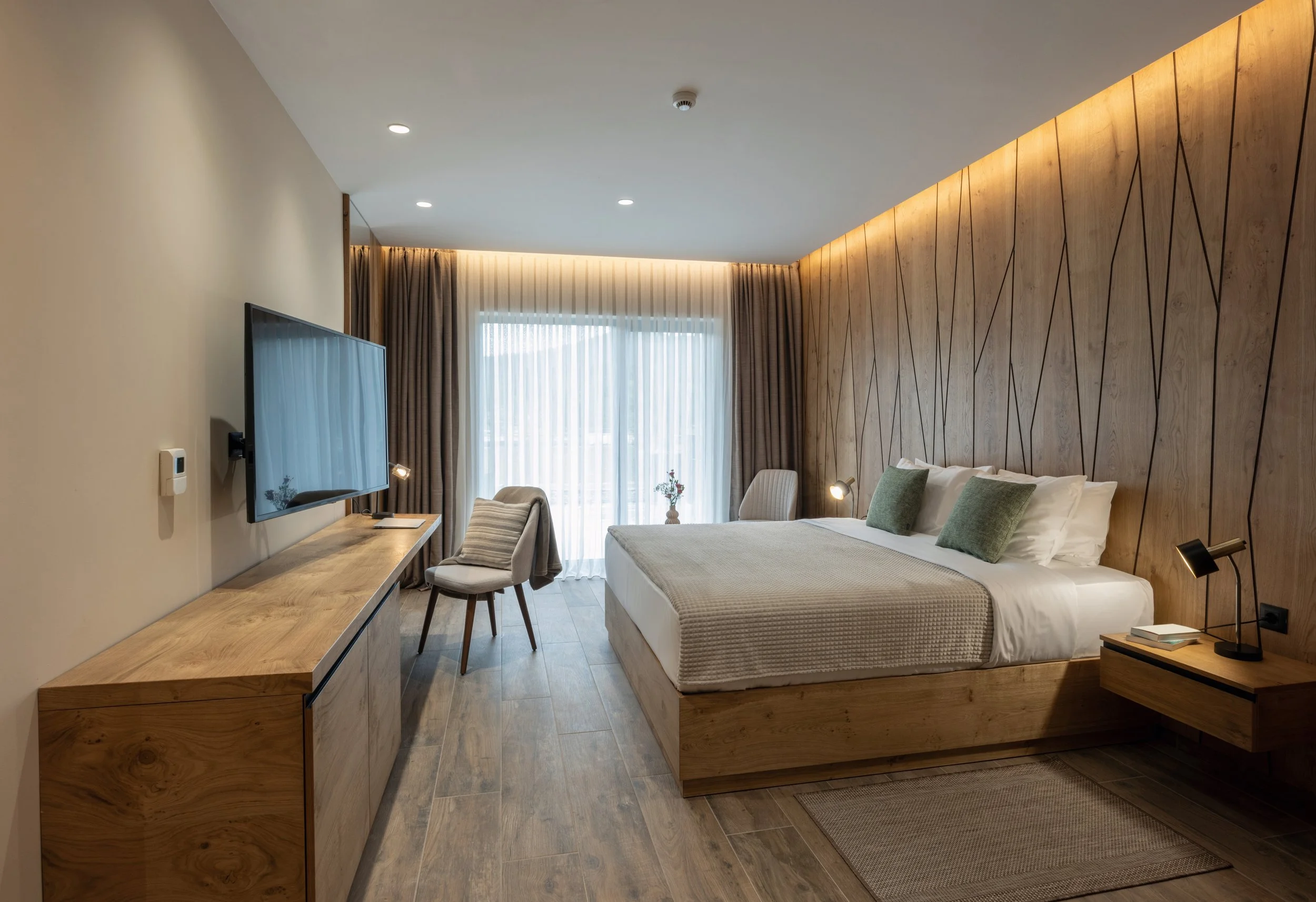 Modern hotel room with a large bed, wooden accent wall, flat-screen TV, and a window with curtains.