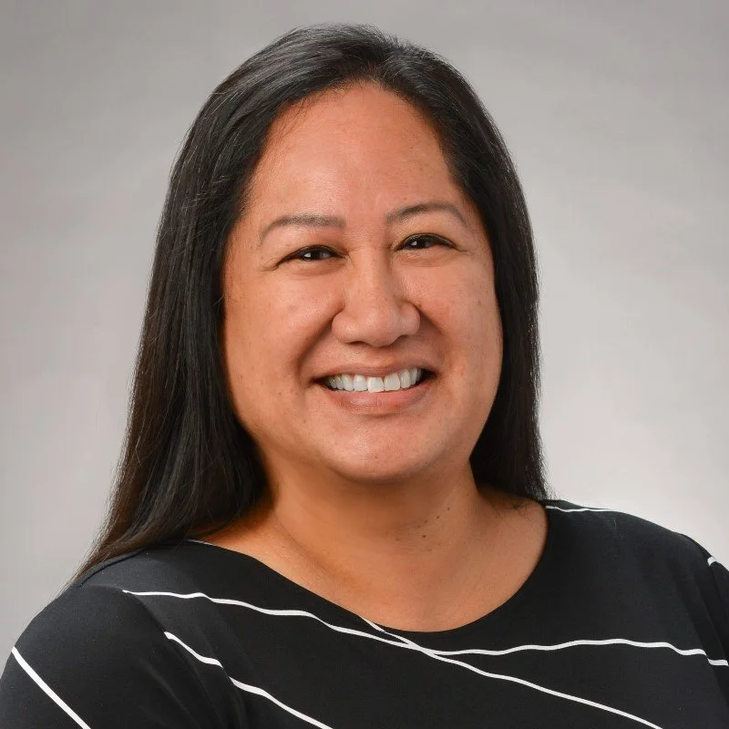 A woman with long black hair smiling at the camera, wearing a black shirt with white lines, against a plain light gray background.