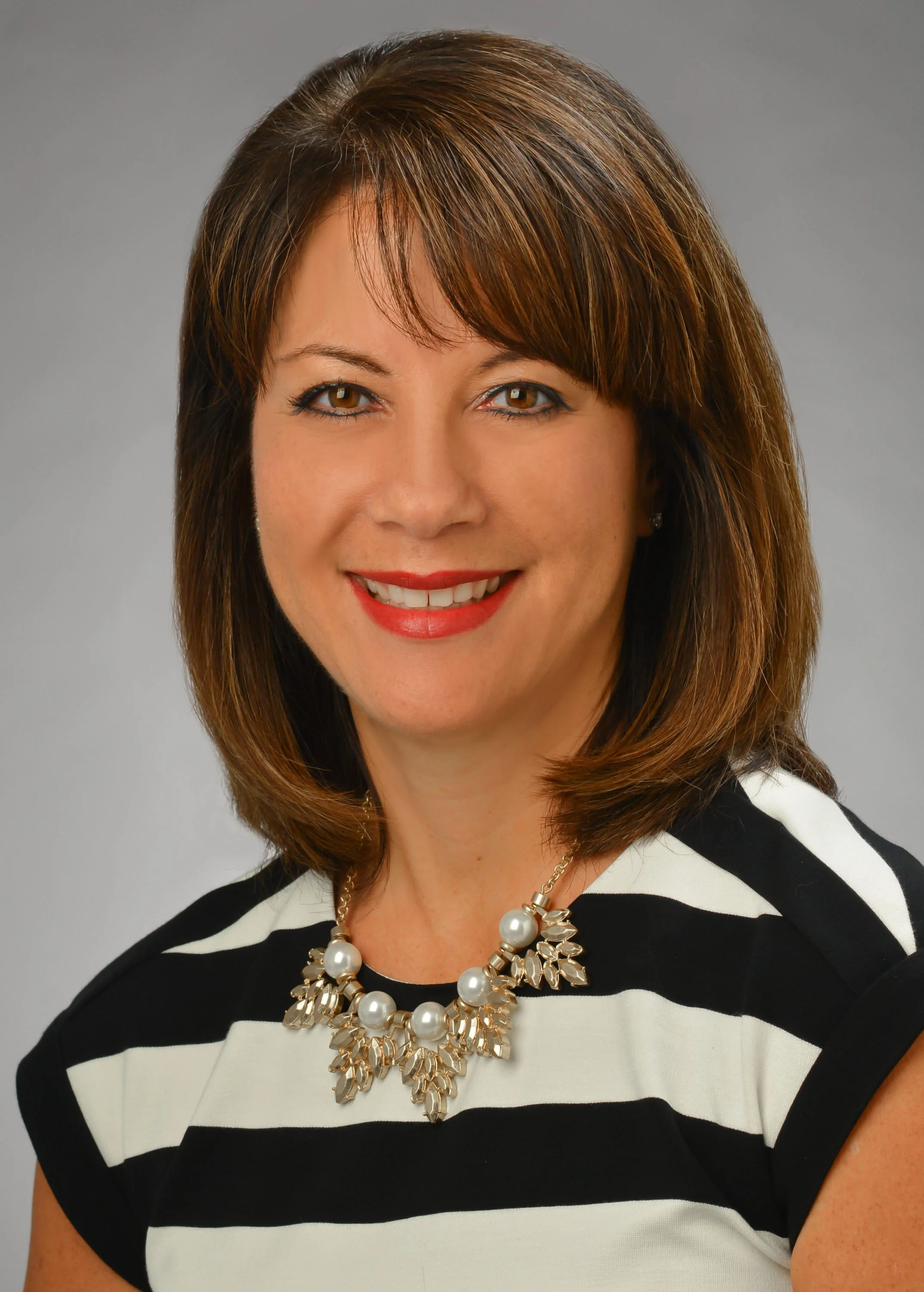 Headshot of a woman with shoulder-length brown hair, wearing a black and white striped top and a pearl and gold leaf necklace, smiling against a gray background.