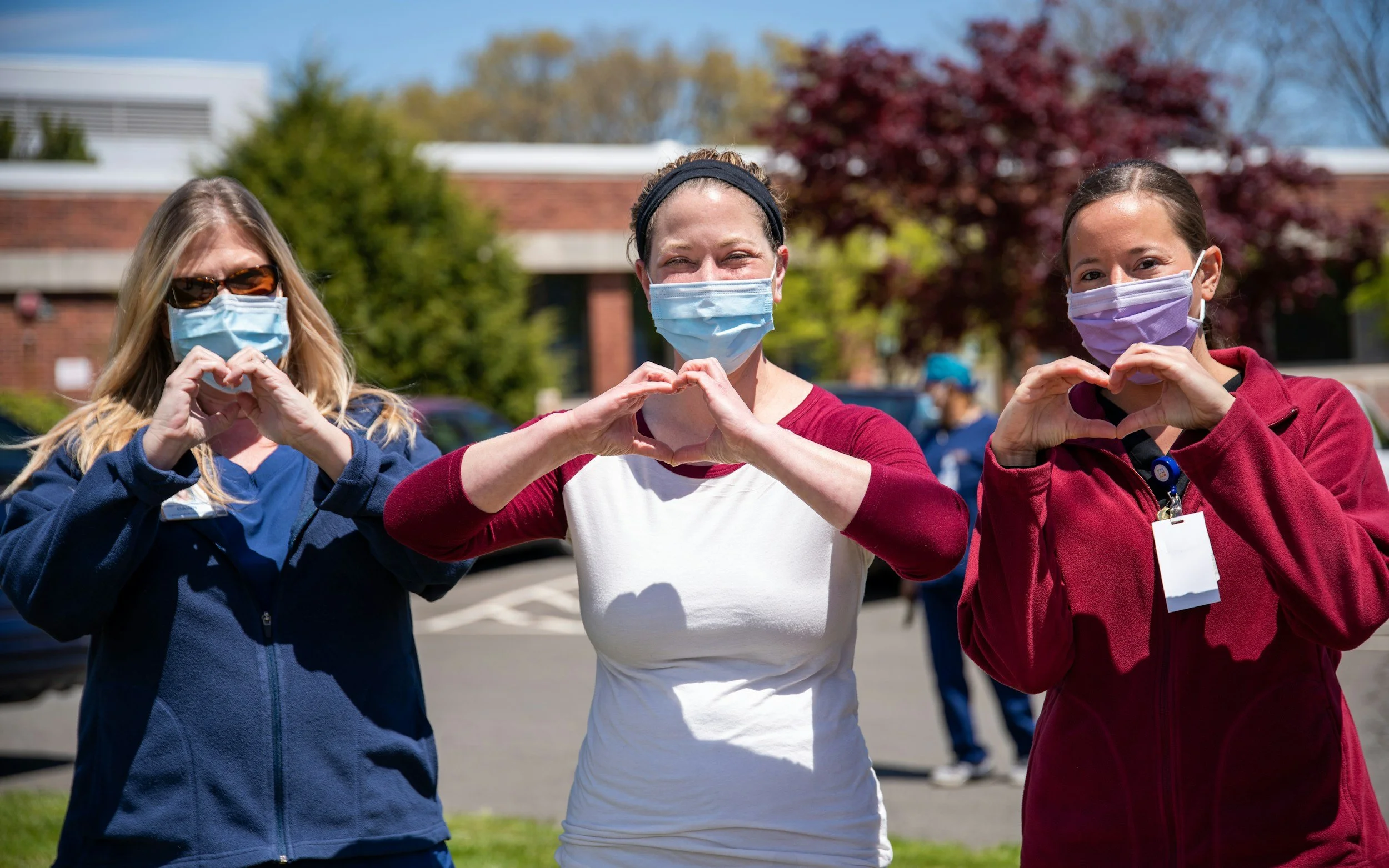 Three women standing outdoors, wearing face masks and making heart signs with their hands, smiling behind their masks.