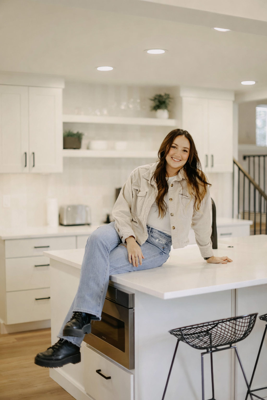 A Bend, Oregon Realtor with long brown hair, wearing a light-colored jacket, white top, and blue jeans, sitting on a modern kitchen island and smiling at the camera at a home managed by the Bend Concierge service.