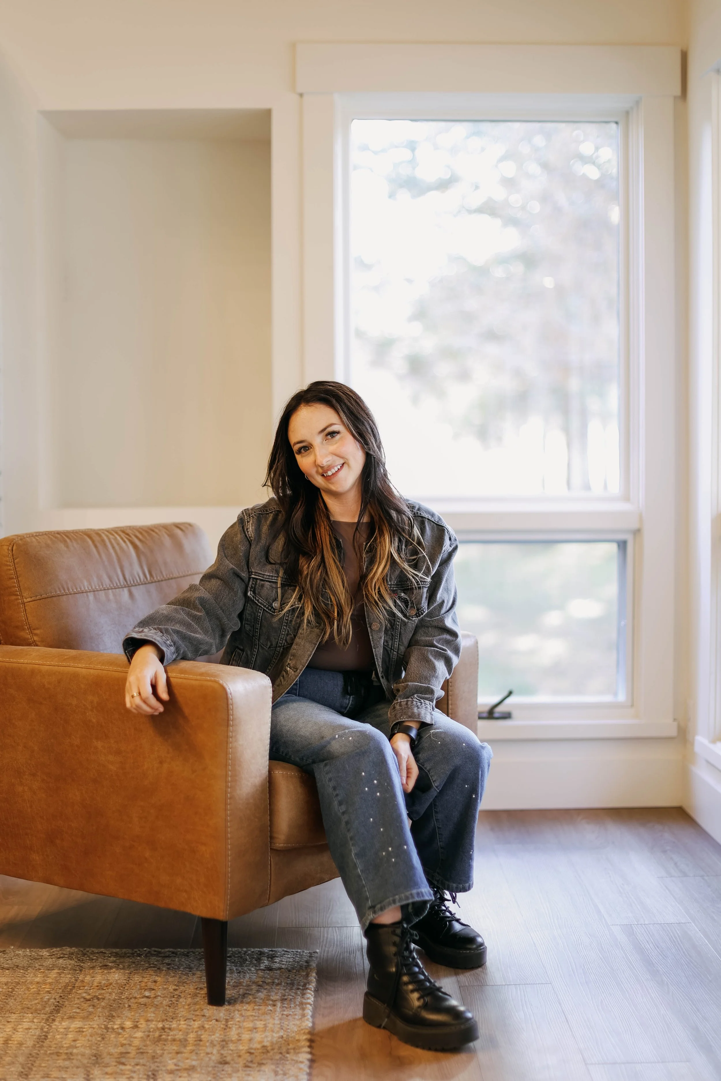 A Central Oregon real estate agent is sitting on a tan leather couch in front of a large window with a view of trees outside in Tumalo, Oregon smiling at the camera. This is a Short-term rental owned by her clients.