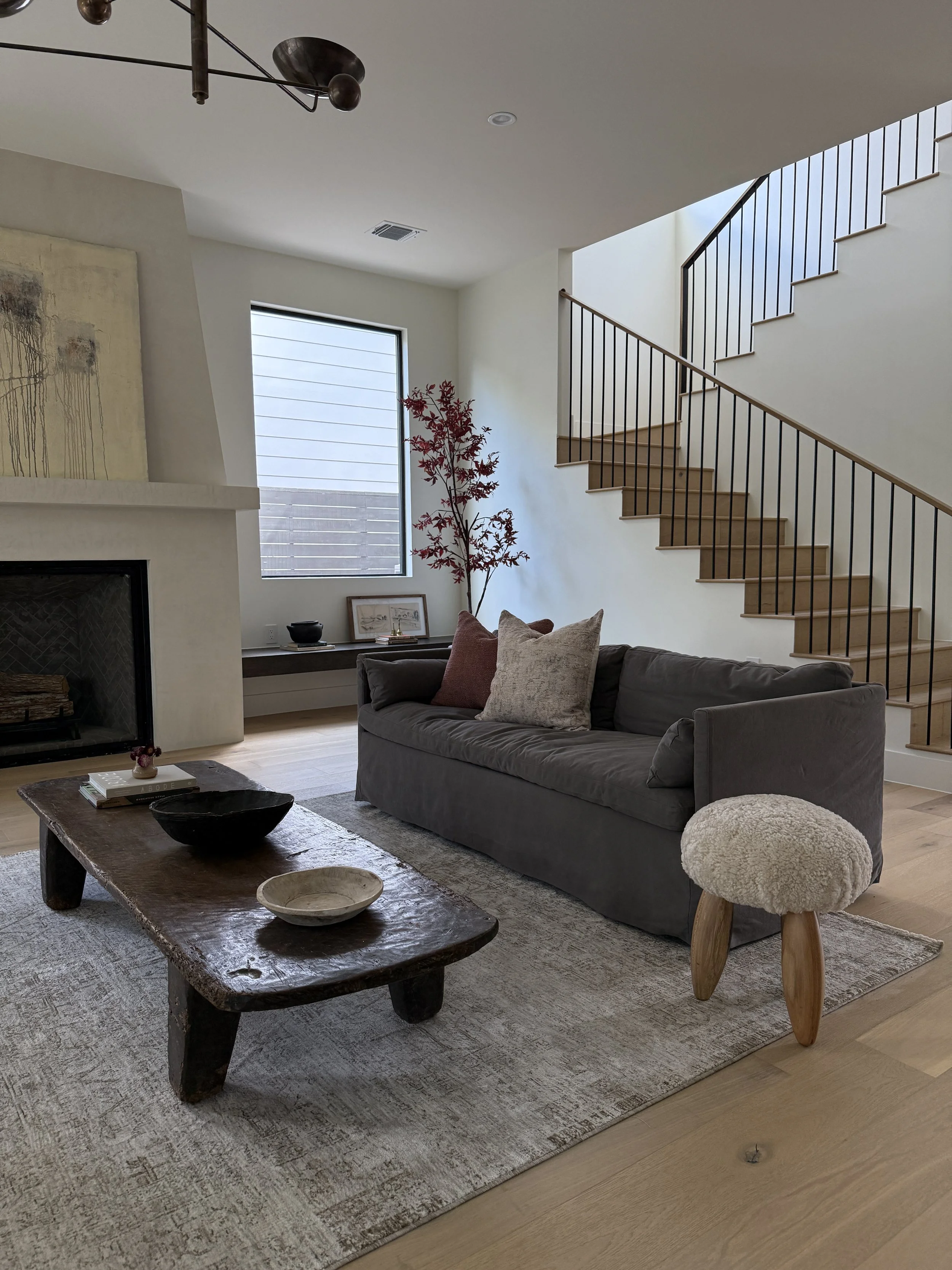Central Oregon Home living room with a gray couch, wooden coffee table, cream-colored ottoman with wooden legs, fireplace, large window, and staircase with wooden steps and black metal railing.