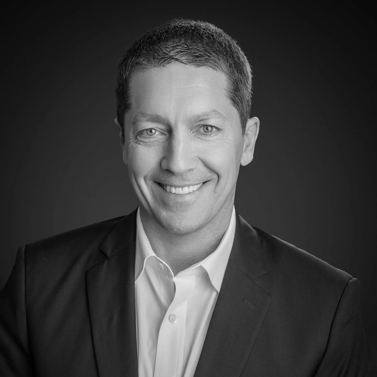 Black and white professional portrait of a smiling man in a suit and white shirt.