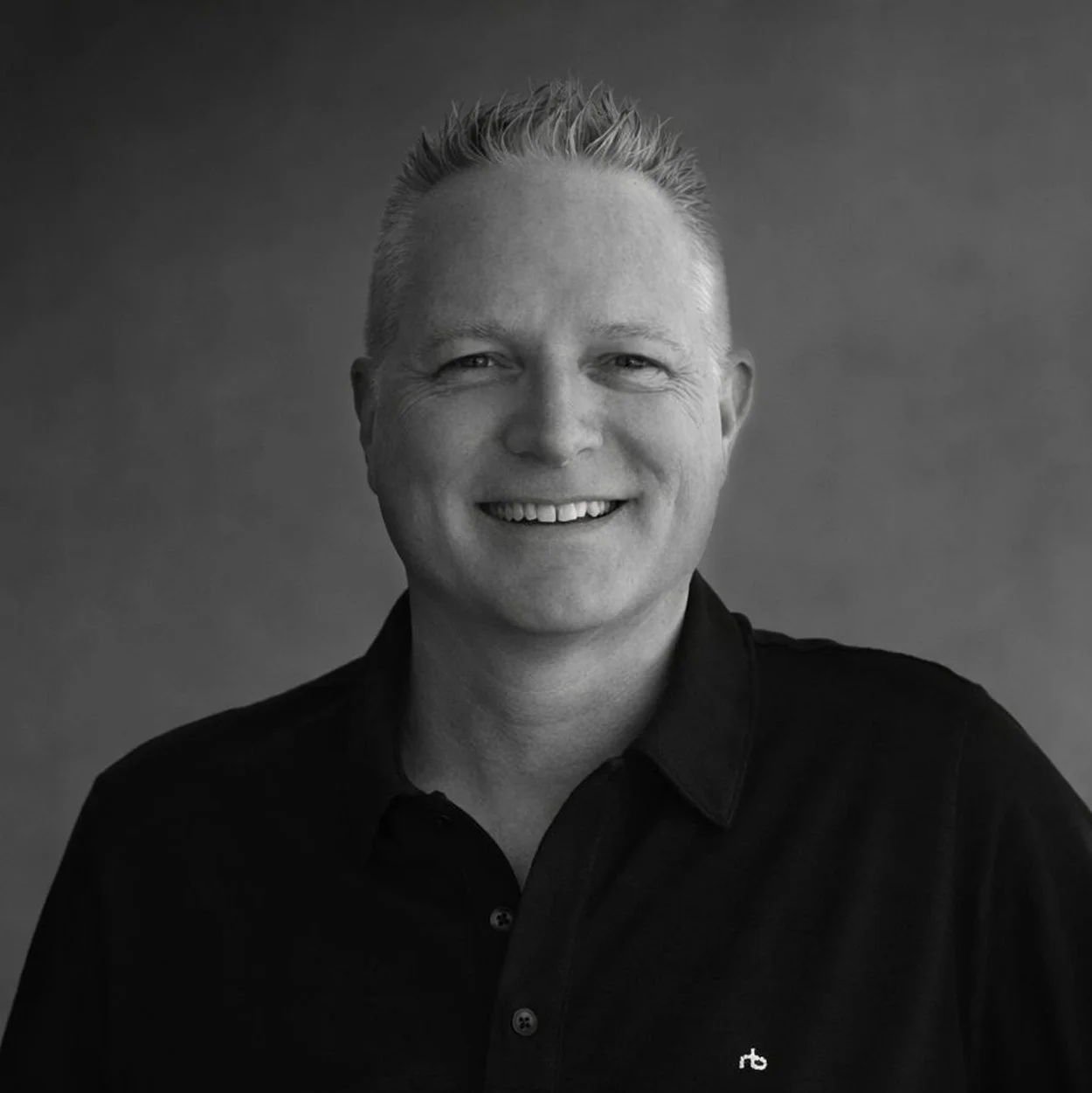 Black-and-white portrait of a smiling man with short, spiky hair, wearing a dark collared shirt against a plain background.