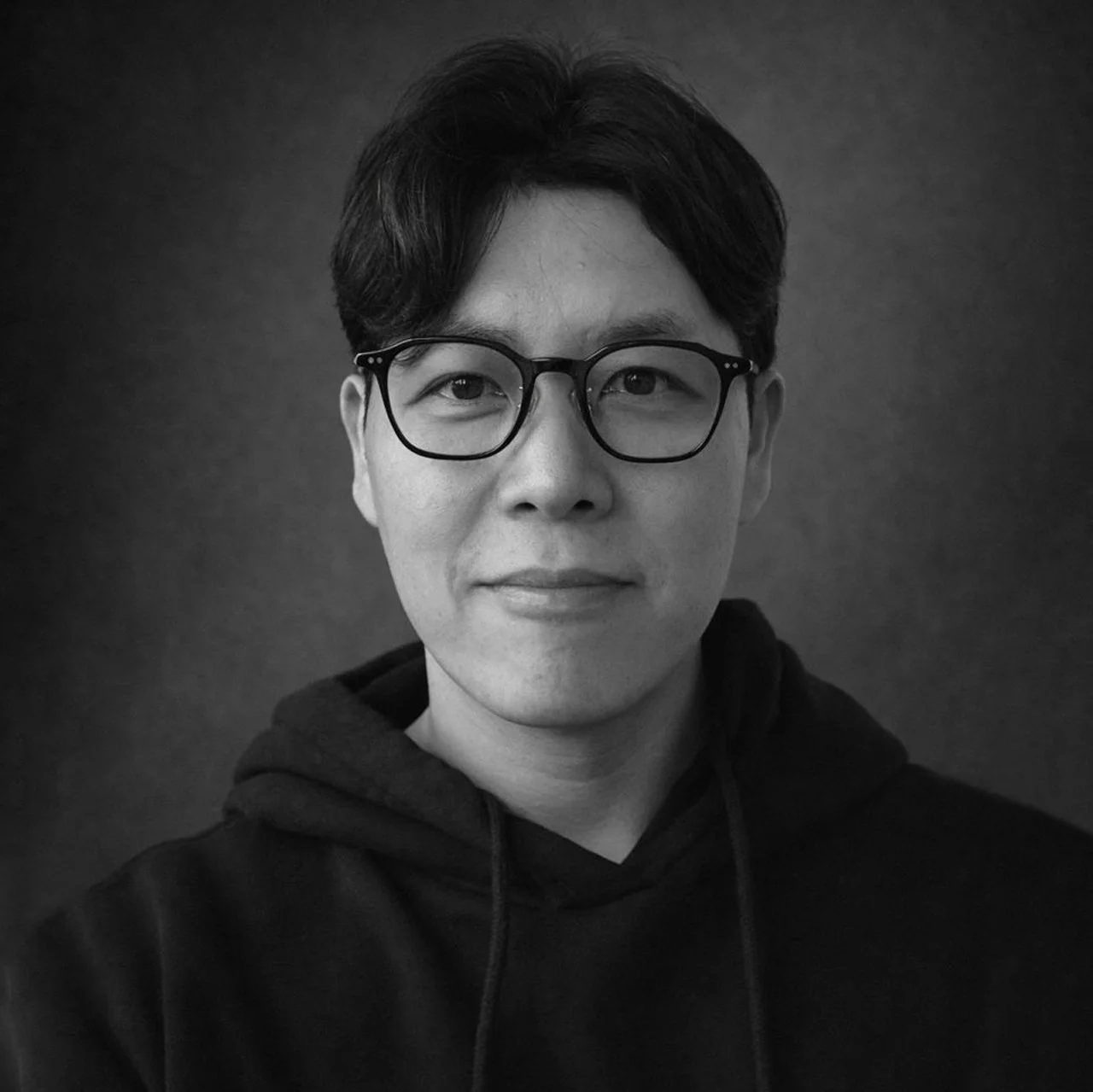 A black-and-white portrait of a man with glasses, short dark hair, wearing a hoodie, against a plain background.