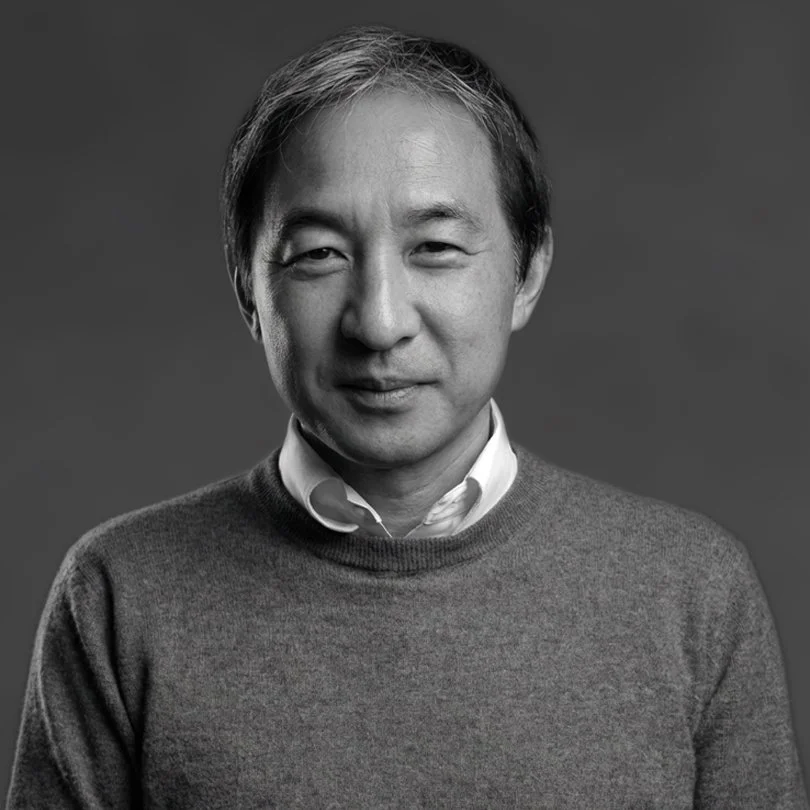 Black and white portrait of a man with short hair, wearing a white collared shirt under a gray sweater, smiling slightly against a plain background.