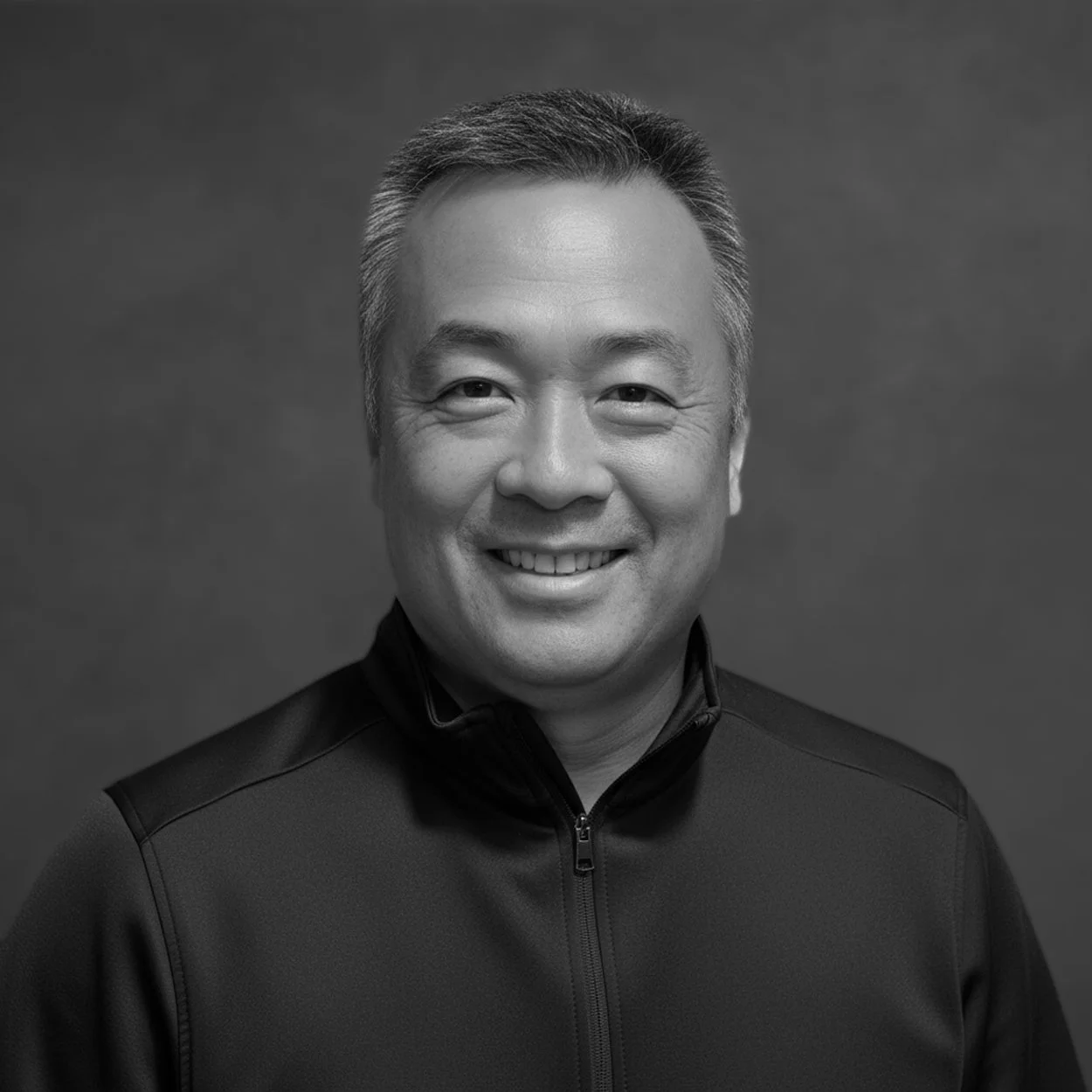 Black and white portrait of a smiling man with short hair, wearing a zip-up jacket, against a plain background.