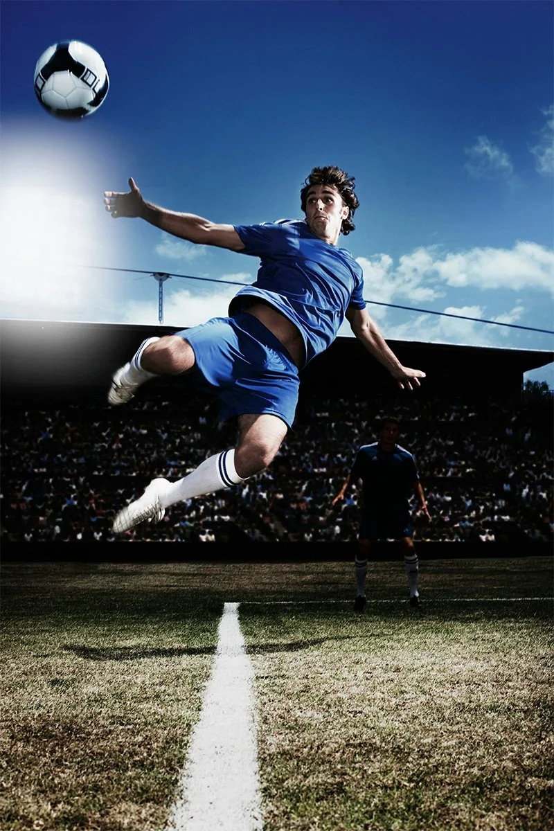 A soccer player in a blue licensed uniform mid-air with a soccer ball during a game on a stadium field.