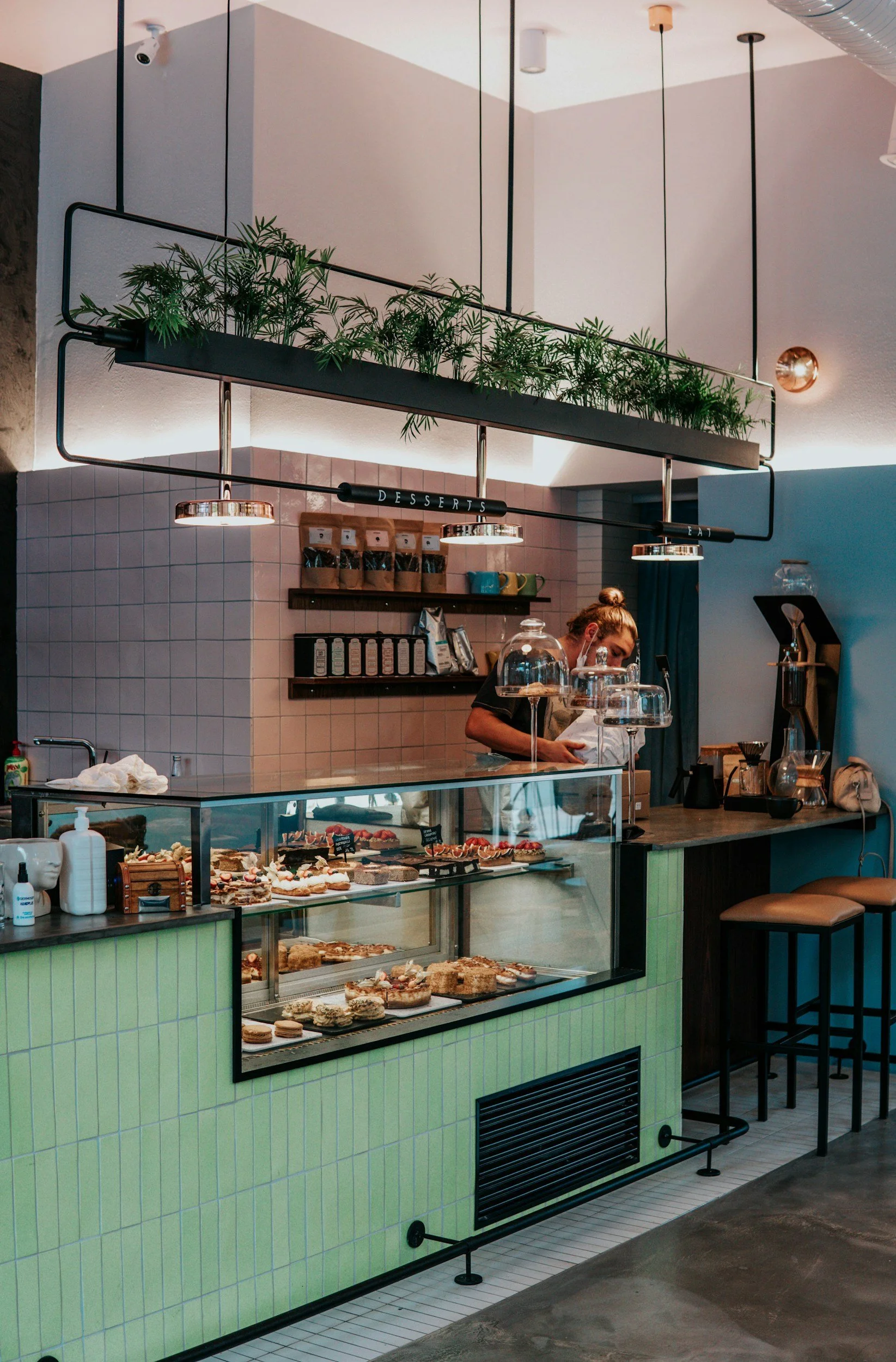 Interior of a modern cafe with a display case of assorted pastries, a barista working behind the counter, and a hanging metal shelf with potted plants. There are bar stools along a counter on the right.