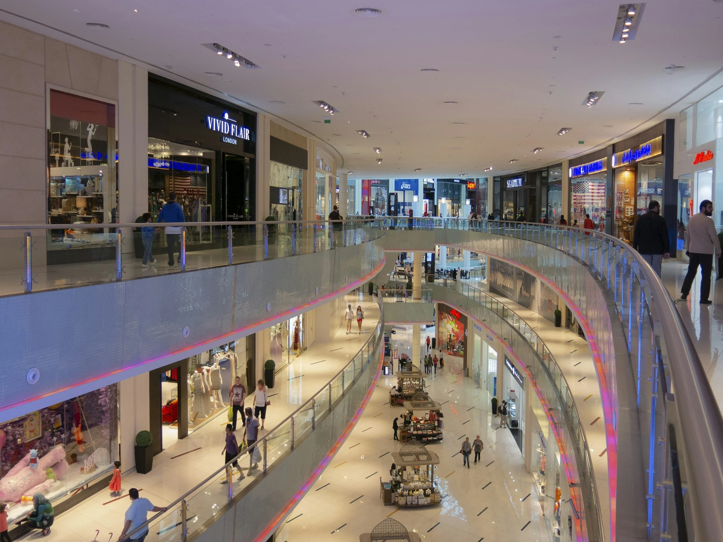 Interior view of a multi-level shopping mall with stores, shoppers, and colorful lighting.