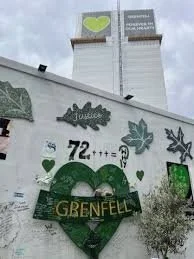 Graffiti-covered wall with a green heart and the word "Grenfell" inside it, surrounded by various painted leaves and symbols, under a cloudy sky.