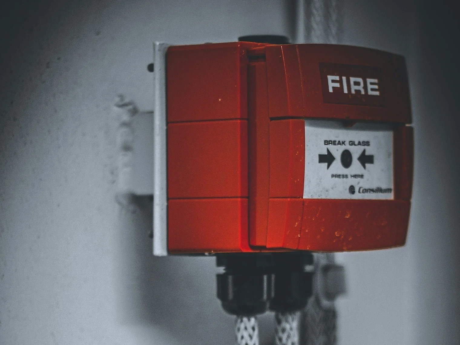 Red fire alarm pull station mounted on a gray wall with a white label and black arrows.