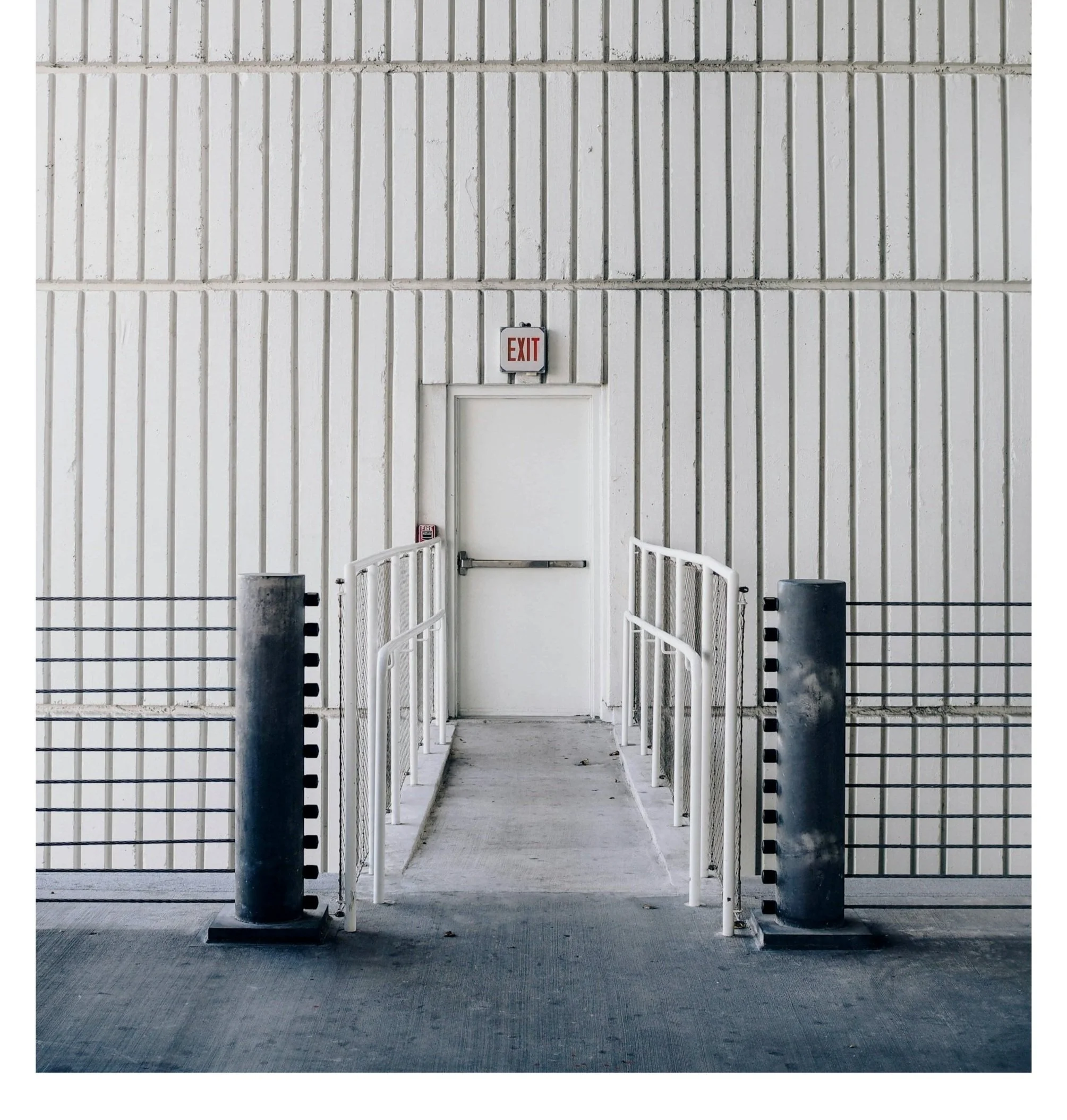 White door with an exit sign above it, accessible ramp with railings on both sides, and safety posts at the entrance, set against a textured white wall.