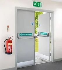 An open fire exit door with an emergency exit sign above, a fire extinguisher mounted on the wall to the left, and emergency instructions on the door handle. Outside, greenery is visible.