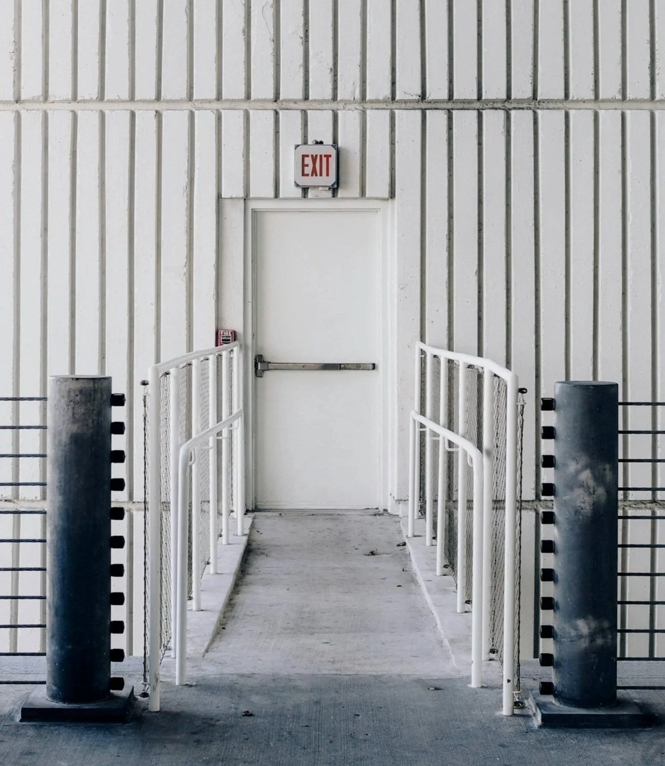 White exit door with an 'EXIT' sign above, accessible ramp and safety rails, set in a plain white wall.