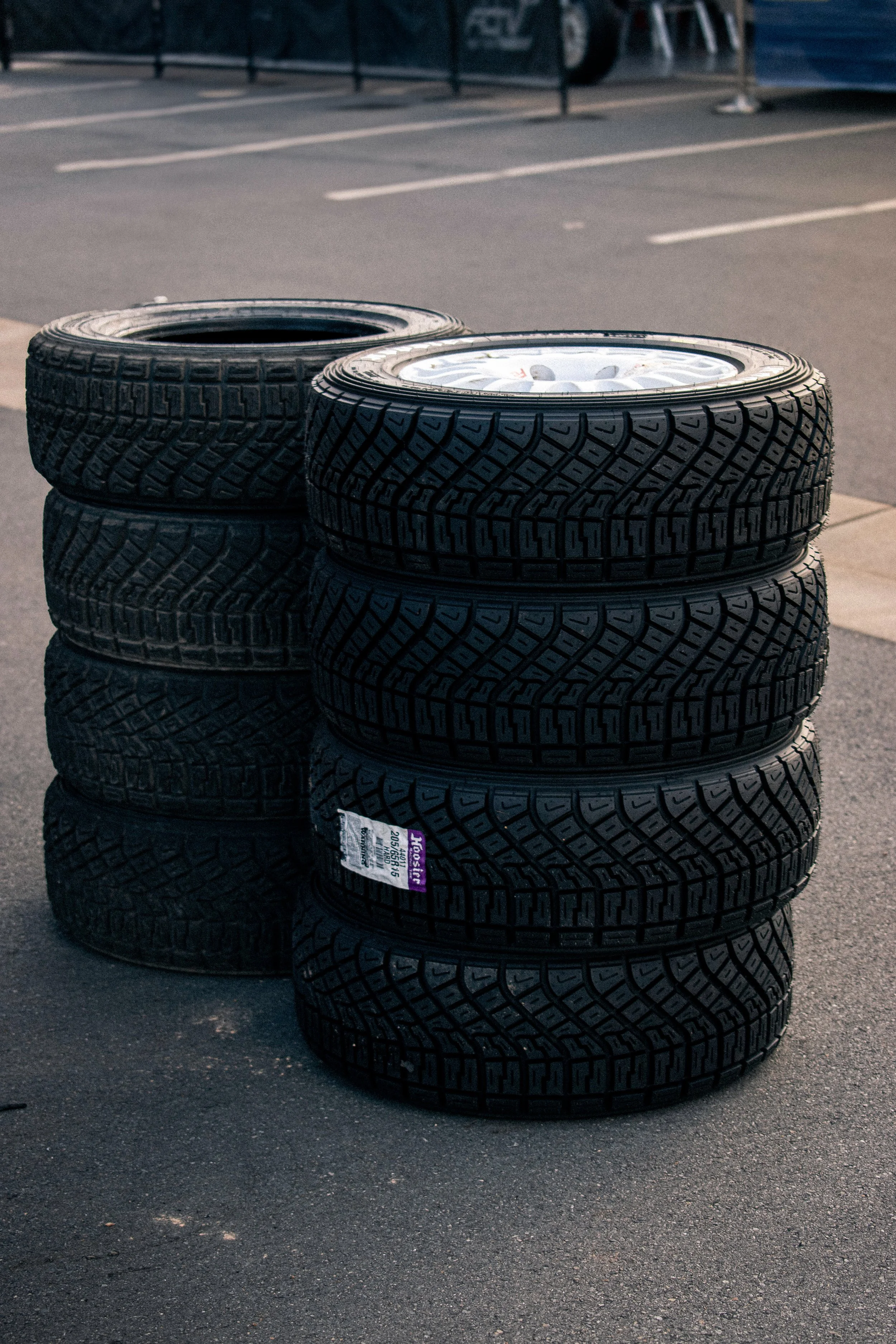 Stack of eight new tires with rims placed on a concrete parking lot near a parking space.