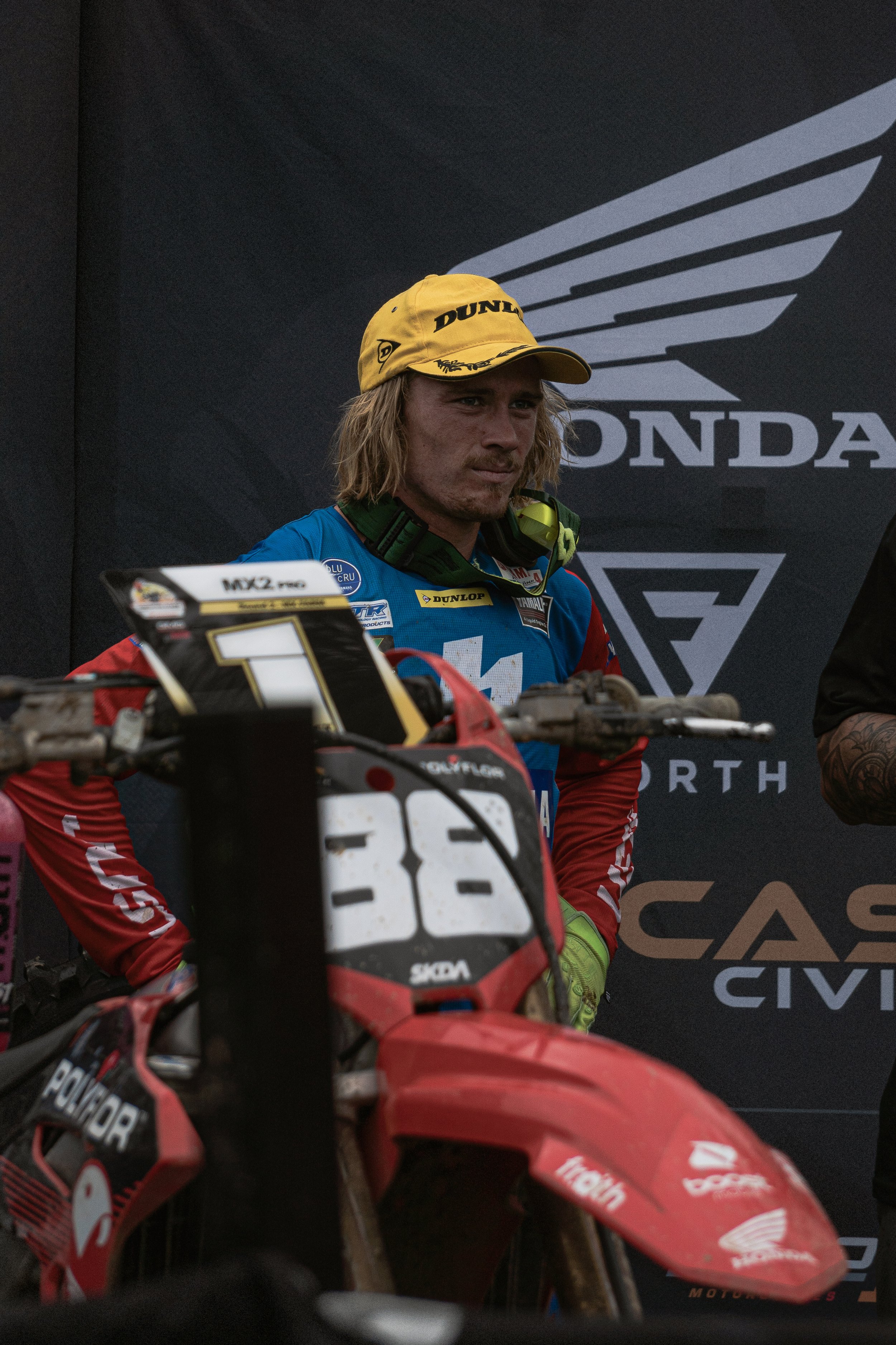 A young man in a blue racing suit with a yellow cap standing next to a red dirt bike, with a black Honda logo background.