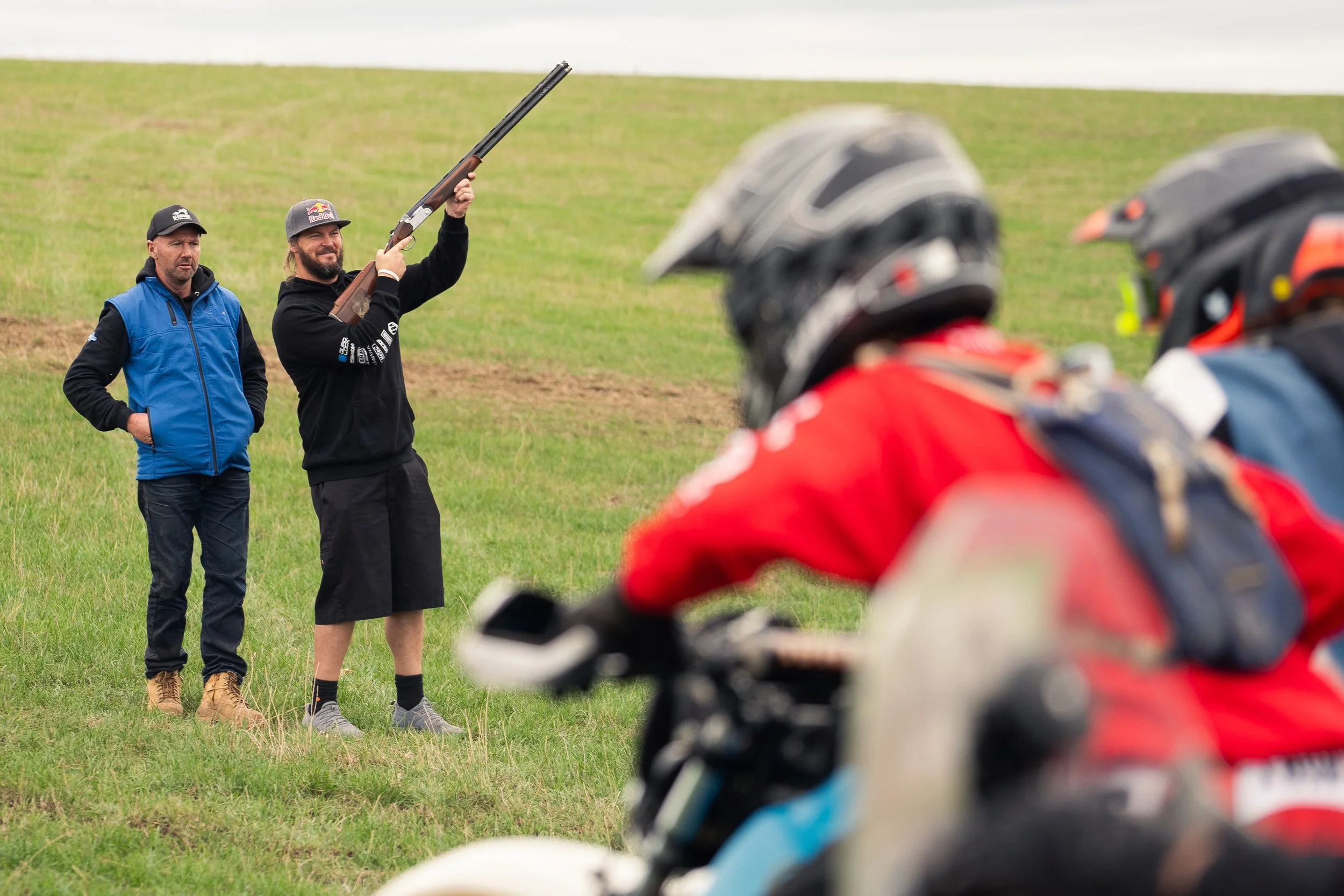 A man with a shotgun raising it in the air while two others watch in a grassy field, with a group of motorcyclists and riders wearing helmets in the foreground.