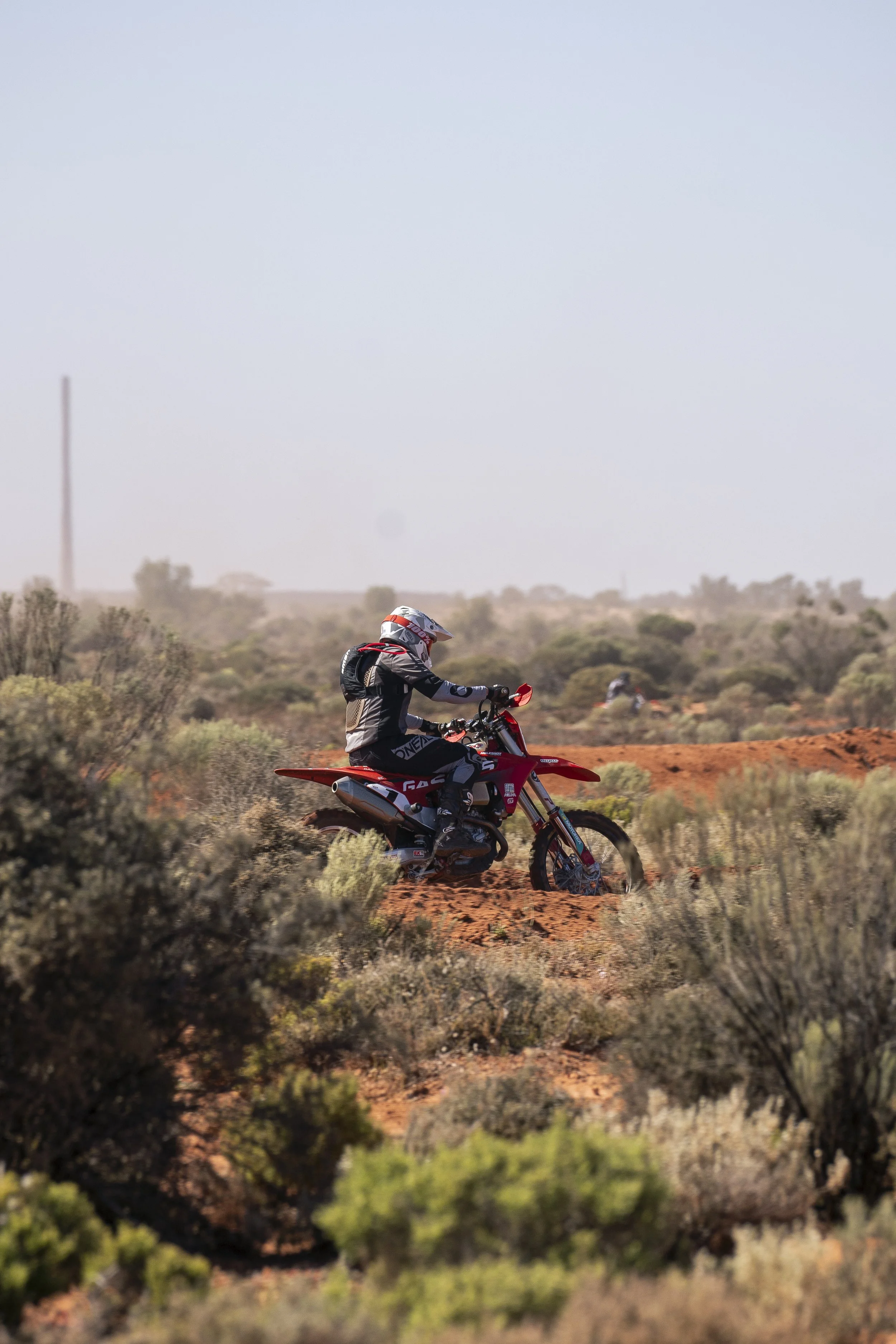A person dressed in motocross gear riding a red dirt bike through a desert landscape with sparse vegetation and a clear sky.