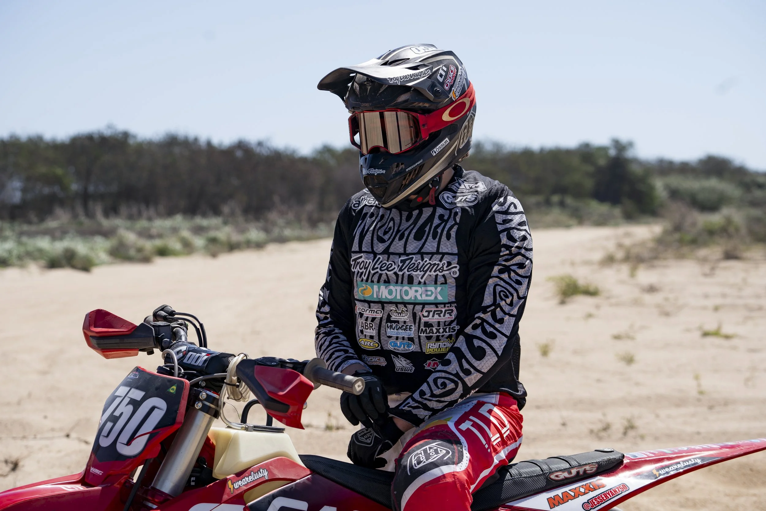 Motocross rider wearing a black helmet with a gold visor and reflective goggles, sitting on a red and white dirt bike in a sandy outdoor environment with sparse vegetation.