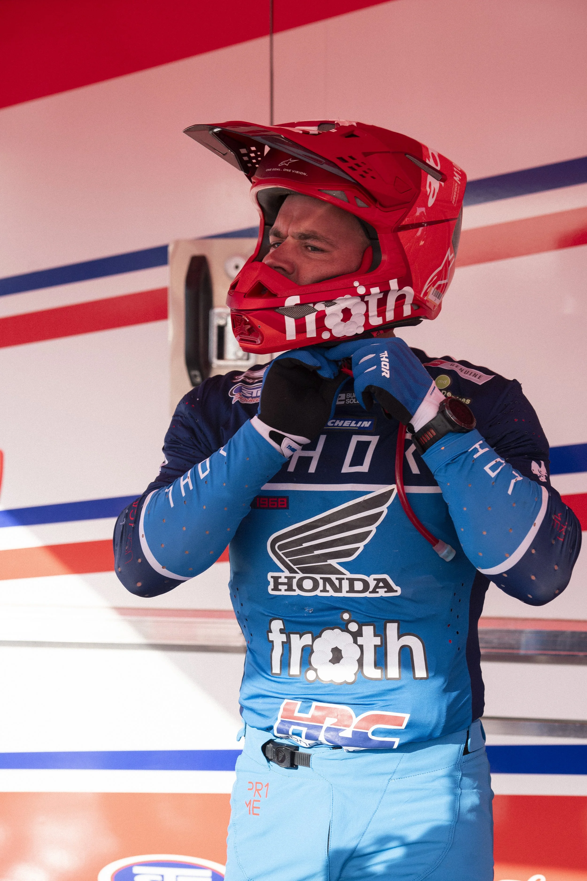 A race car driver wearing a red helmet and blue racing suit, adjusting his gear before a race.