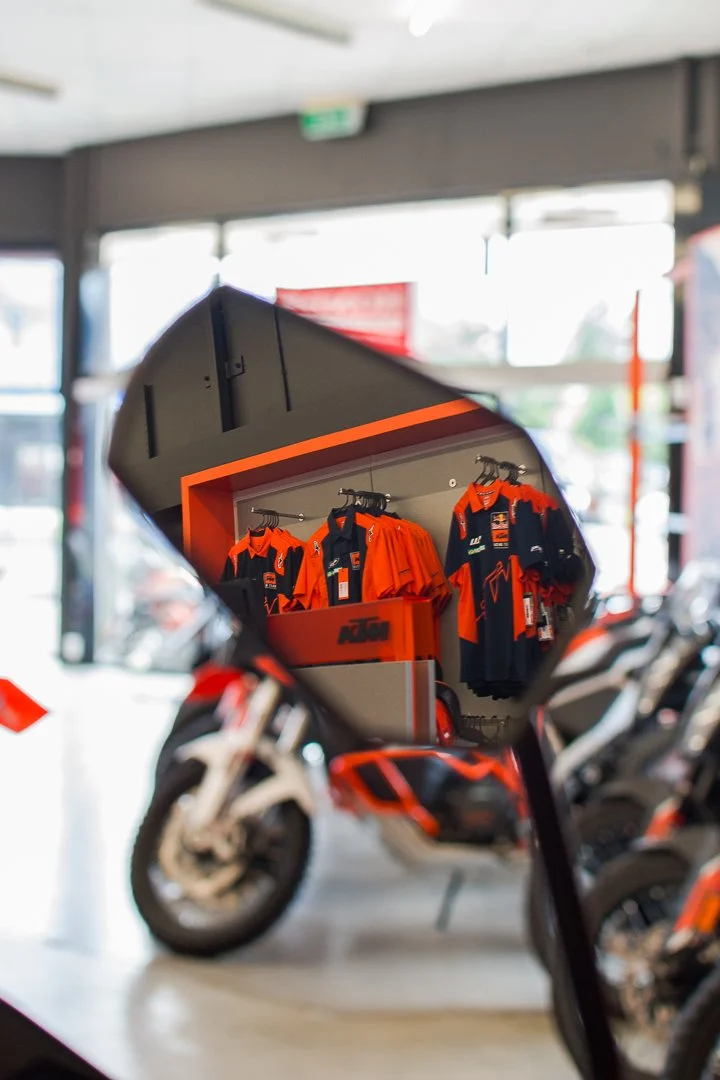 Motorcycles on display inside a dealership, viewed through a mirror reflecting a display of motorcycle apparel.