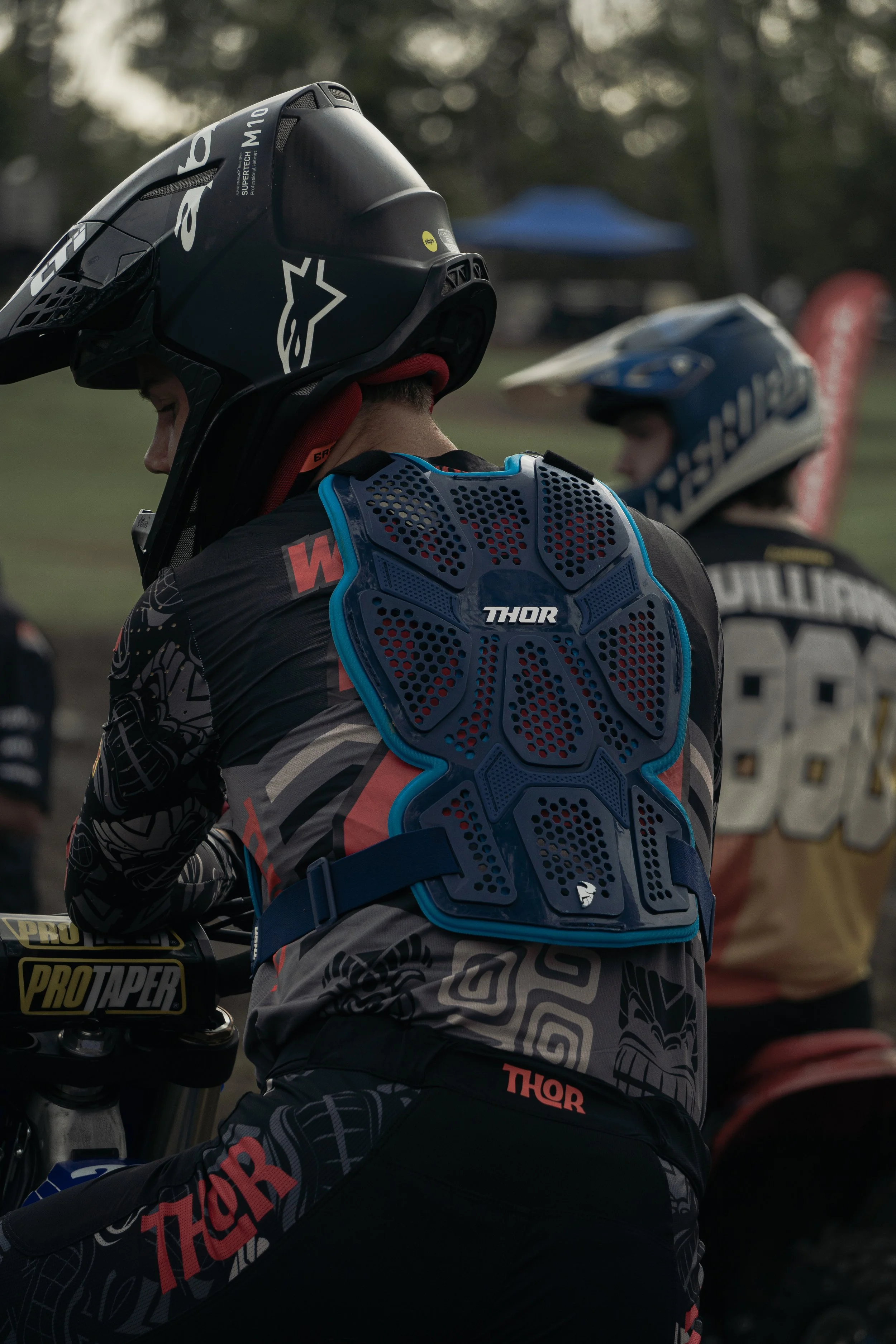 Two motocross riders wearing full gear, including helmets and chest protective gear, preparing for a ride outdoors with trees and tents in the background.