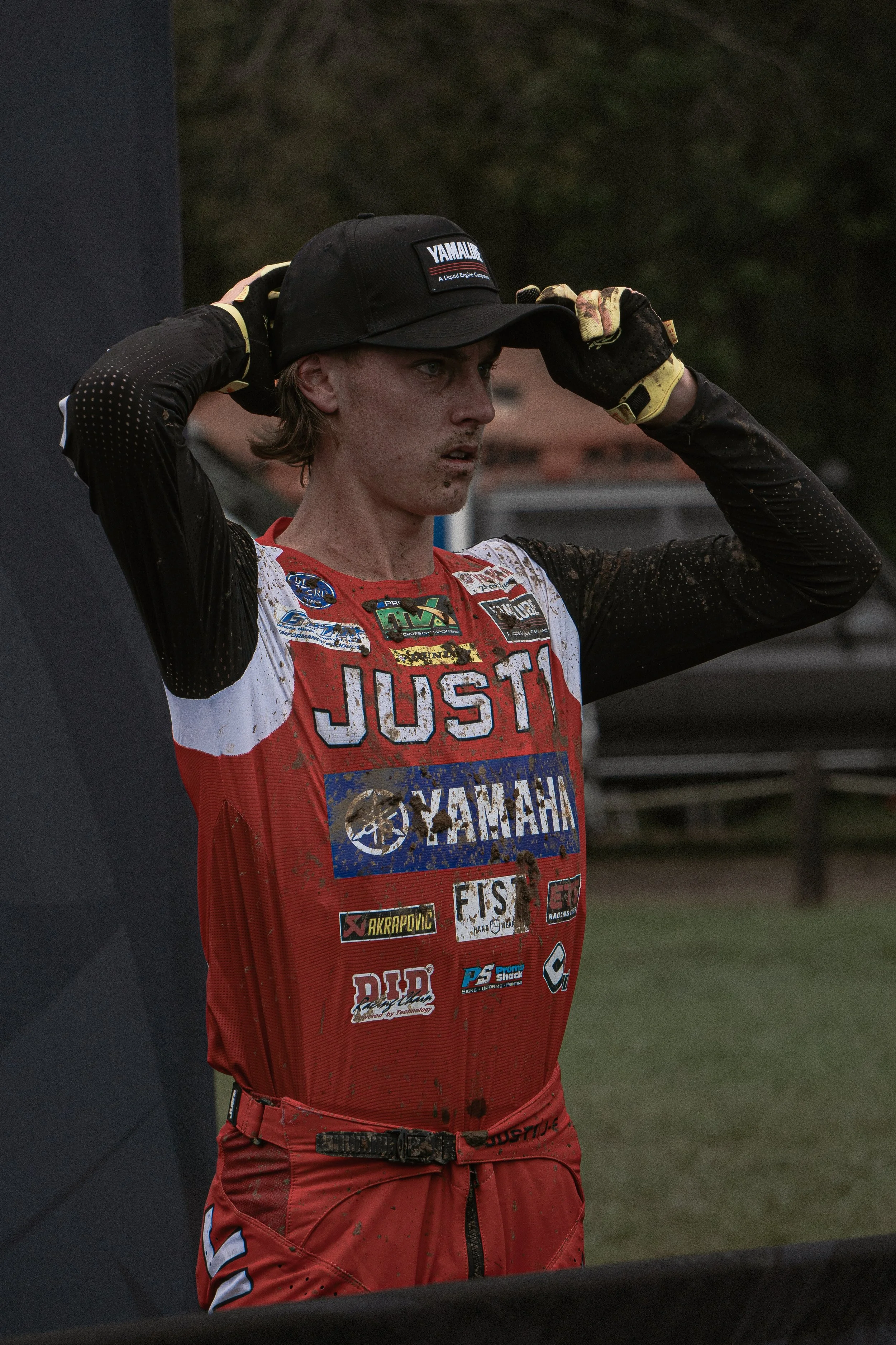 A young motocross racer is adjusting his black hat after a race, wearing a red and white racing jersey with numerous sponsorship patches, with dirt smudges on his face and clothes, outdoors on a dirt track.