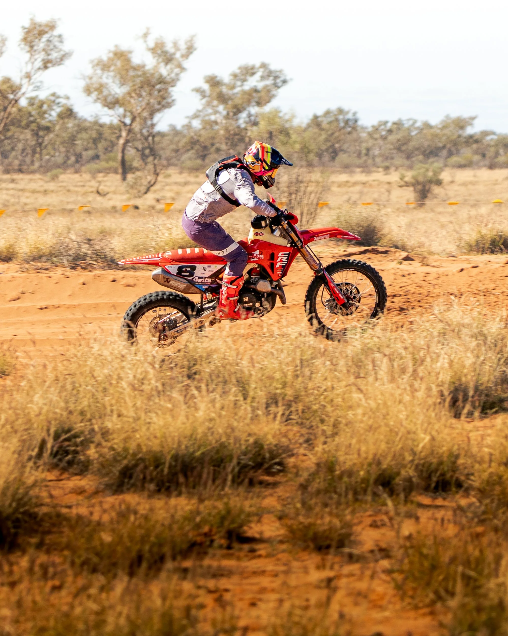 A person riding a red dirt bike through a desert landscape with dry grass and sparse trees in the background.