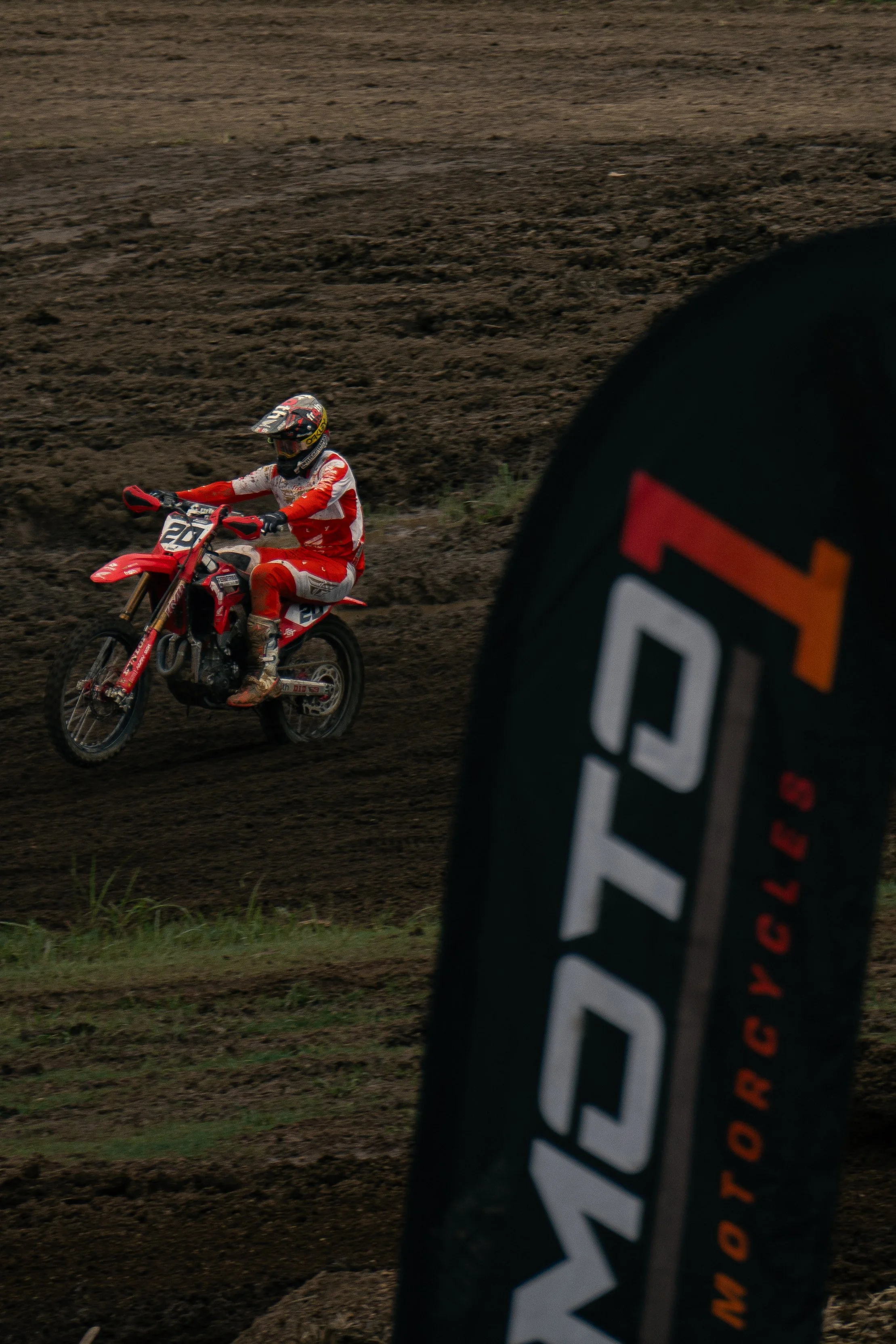 Motocross rider in red and white gear riding a dirt bike on a muddy track, with a partial View of a black and orange motocross wheel in the foreground.