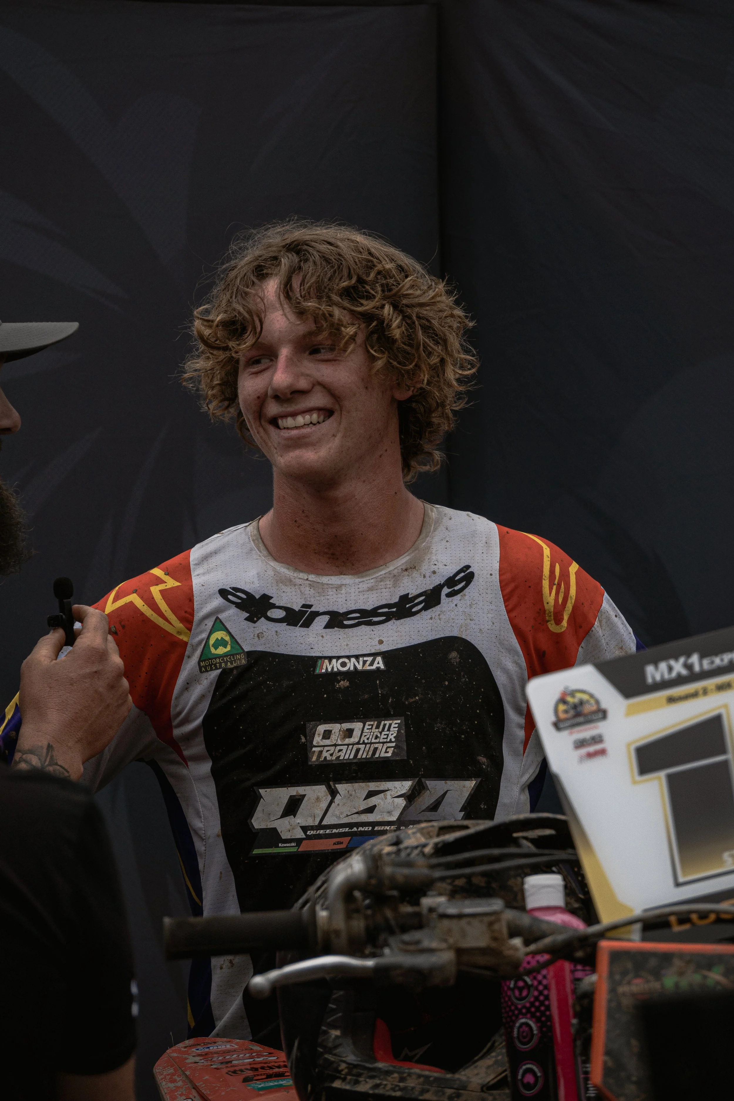 A young male motocross rider with curly hair smiling after a race, standing beside his dirt bike, wearing a dirt-streaked racing jersey with sponsorship logos.