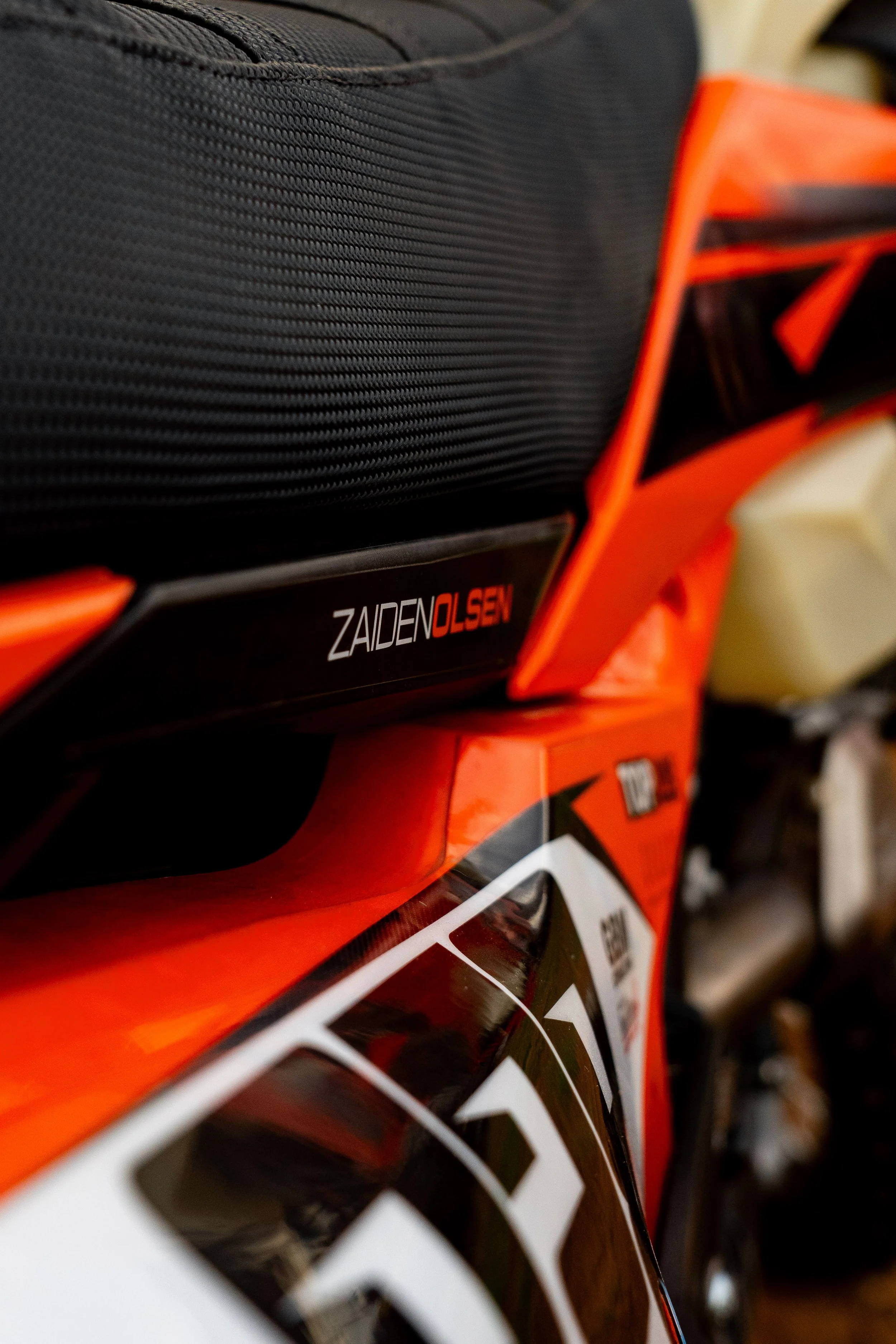 Close-up of a motorcycle with black and orange bodywork, featuring a black luggage compartment and a small sticker with the name 'Zaiden Olsen'.